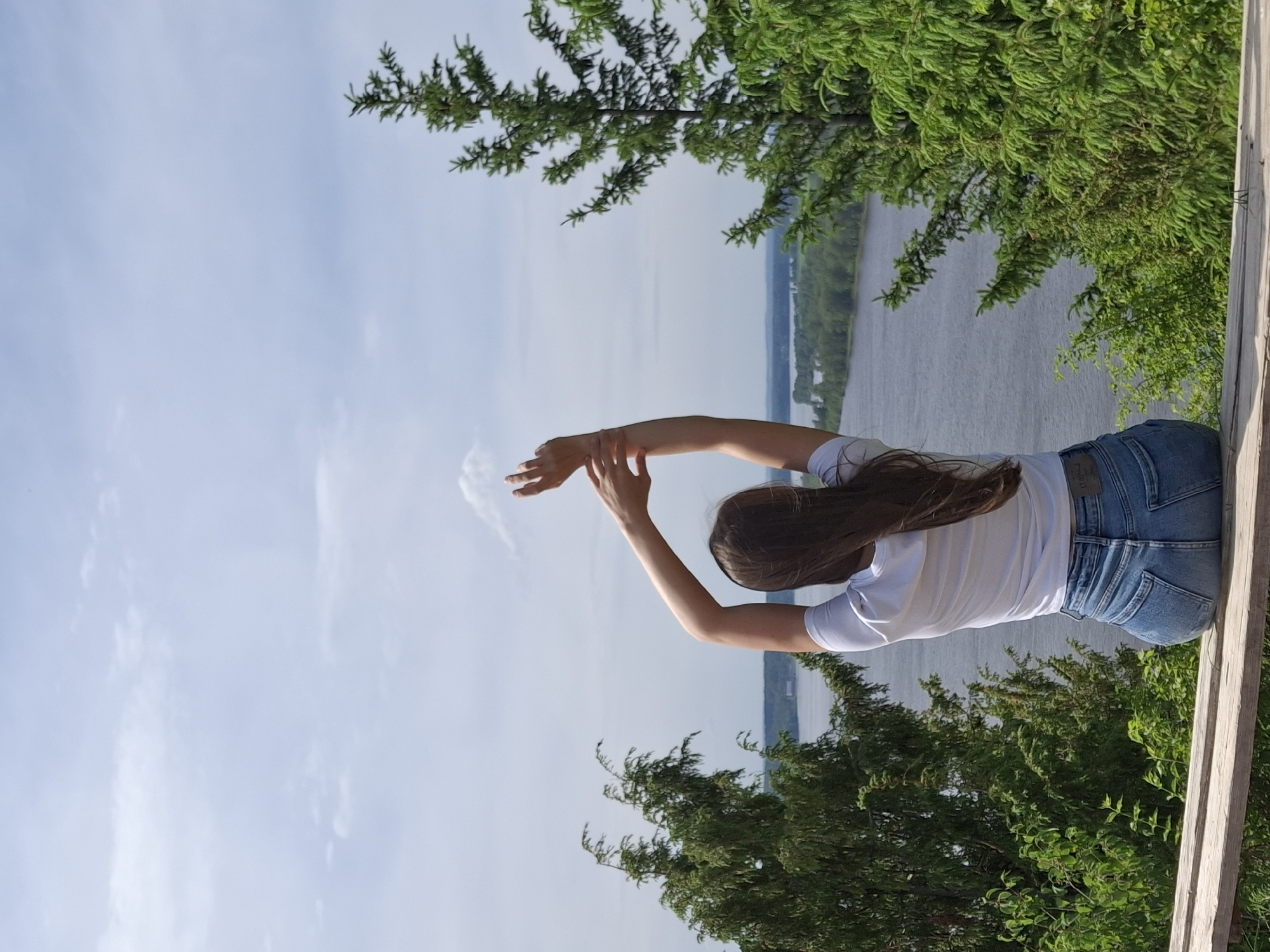 A person stands along a wooden barrier. In the background there is a river with tree lines on the opposite side. The sky is cloudy. This photograph was taken in Ольховка, Russia.