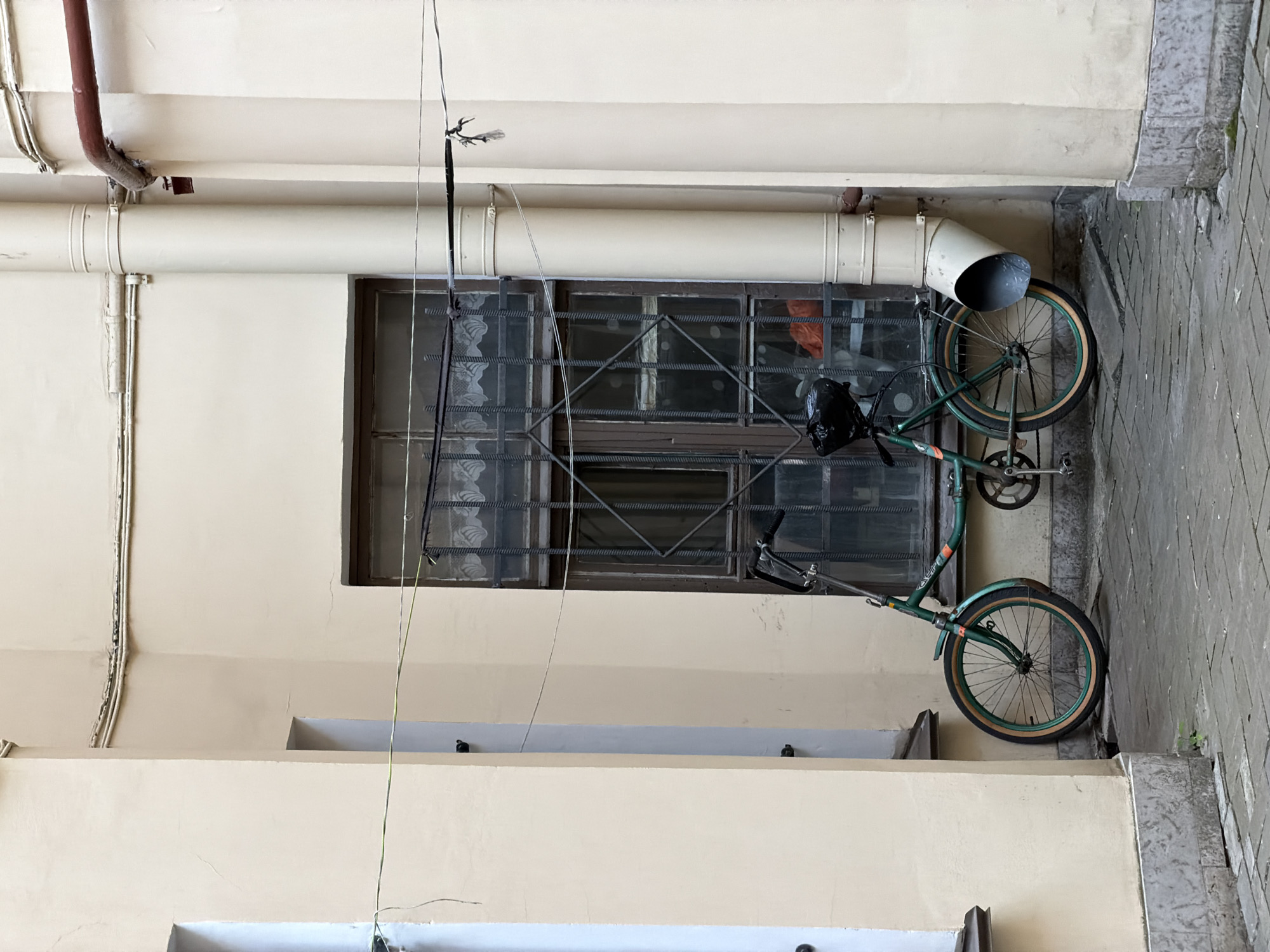 A green bicycle is leaning against a cream-colored building next to a window with a metal grate. The bicycle has a black bag on the handlebars. A large, beige pipe extends from the building above the bicycle and curves downward, with a funnel-shaped opening. The building has a rough texture and appears to be old. The ground is paved with gray bricks. The scene is outdoors, and the lighting suggests it is daytime. The image is oriented vertically, with the top of the bicycle at the bottom of the frame. The overall impression is one of an urban courtyard or alleyway. There are no people visible in the image.