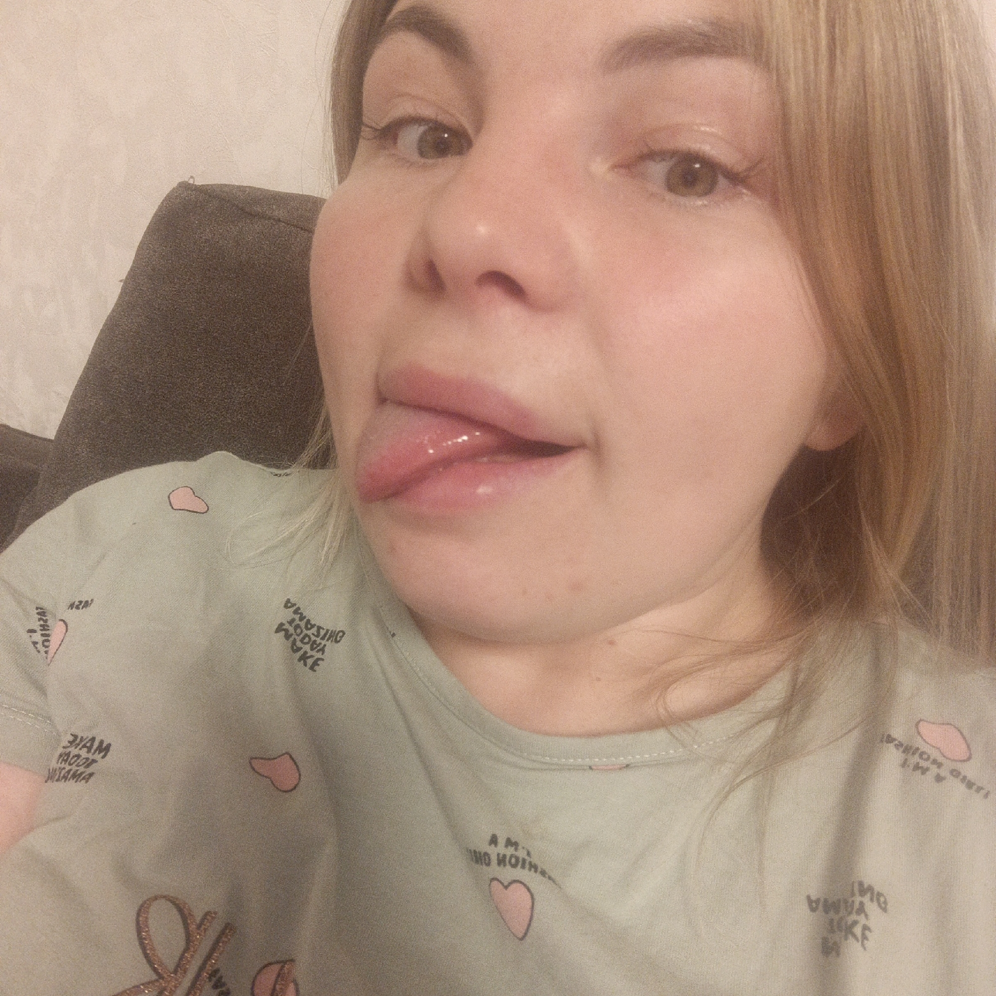 A close-up selfie shows a person, likely a woman, with fair skin and light brown or blonde hair, sticking out their tongue in a playful or silly gesture. Their eyes are open and directed slightly upwards and to the right. The person's lips appear glossy, and the extended tongue is moist.
They are wearing a light sage green t-shirt patterned with small pink heart shapes. Several words are printed on the shirt in reverse, including phrases that appear to be "AM I LOVING THIS" and potentially Japanese words like "OHISAMA" (sun) and "YAMA" (mountain), along with "WAKE".
The background features a dark, possibly grey, sofa or chair arm on the left side, and a textured, light-colored wall behind the subject. The lighting is somewhat dim and artificial, consistent with an indoor setting. The overall mood of the image is informal and lighthearted.