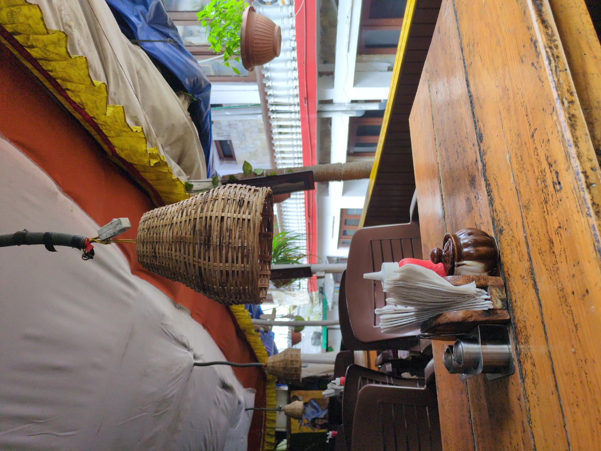 An outdoor dining area is depicted, likely at a cafe or restaurant in Bhagsu, India. The foreground is dominated by a rustic, weathered wooden table. On the table, a set of stainless steel salt and pepper shakers, a wooden holder with folded white napkins, a red squeeze bottle of ketchup, and a small, lidded ceramic jar are arranged. In the background, brown plastic chairs are visible, set at tables. Above the dining area, a white fabric canopy is stretched taut, with an orange fabric layer and decorative yellow and blue trim visible underneath, suggesting an awning or shelter. A woven bamboo lampshade hangs from a black electrical cord near the center of the frame. Further in the background, a white railing lines a balcony or walkway, with buildings and greenery beyond. The scene conveys a casual, open-air dining atmosphere typical of a popular tourist spot like Bhagsu.