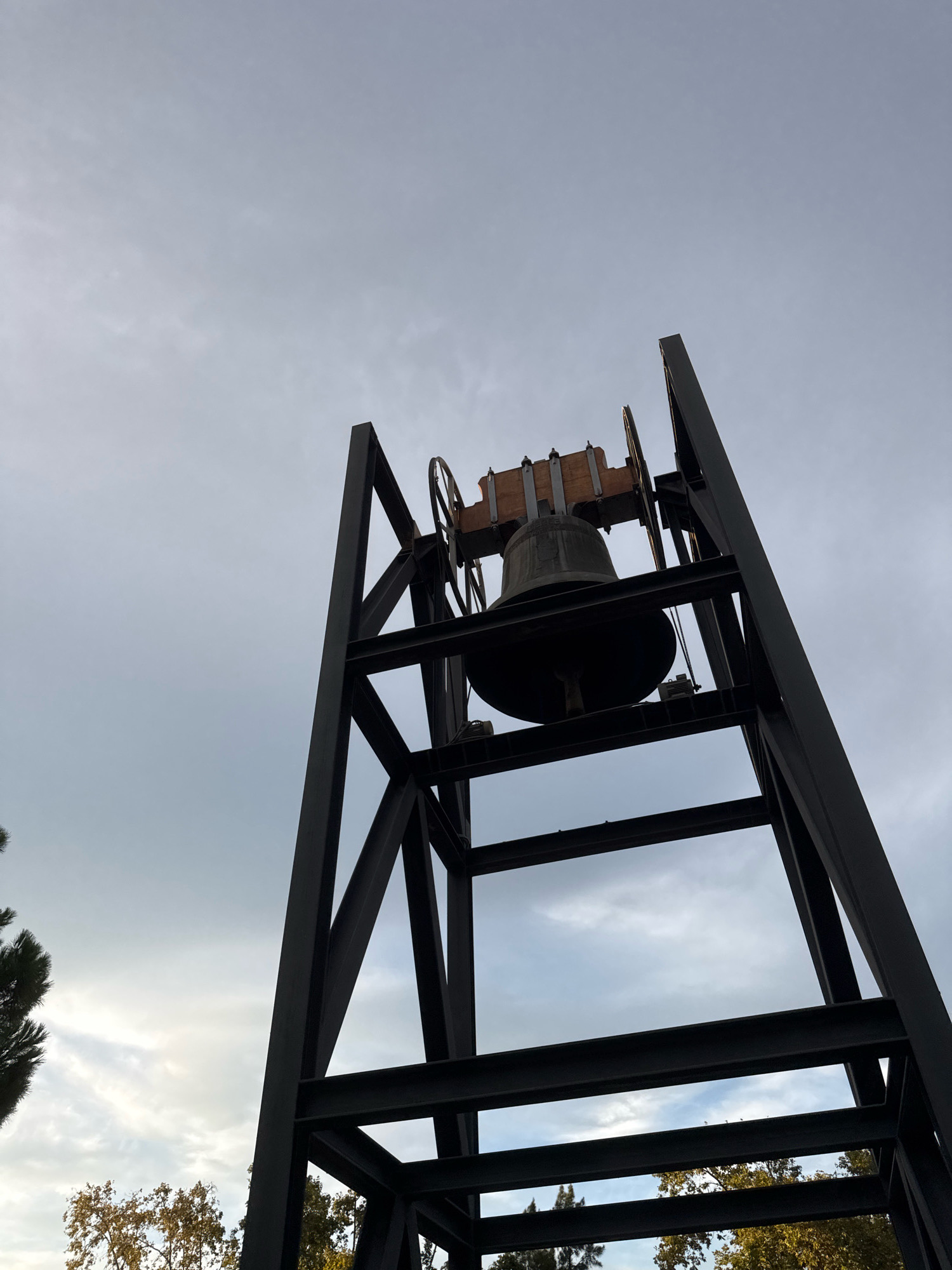 A large, dark bell is suspended from a metal A-frame tower against a cloudy sky. The bell is mounted on a wooden beam with two circular pulleys on either side. The tower is constructed of dark, rectangular metal beams forming a rigid structure. The perspective is from a low angle, looking up at the bell and tower. Trees with some autumnal foliage are visible at the bottom of the frame. This scene is located at the Park Güell in Barcelona, Spain.