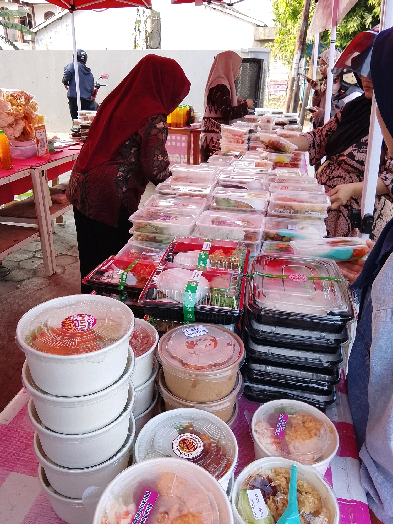 The image depicts an outdoor food stall in Kudaile, Indonesia. A woman wearing a red hijab and patterned blouse is standing behind a table laden with various food containers. She is interacting with the food. In the background, another woman in a beige hijab is visible, also near the food stall. To the left, a person on a motorcycle with a helmet is also present. The stall is set up under a red canopy, with a pink and white checkered cloth covering the table. Numerous plastic containers of food, some stacked, are arranged on the table. Some containers have labels, including one with "Nasi Ayam Acam Manis" visible. The ground appears to be paved.