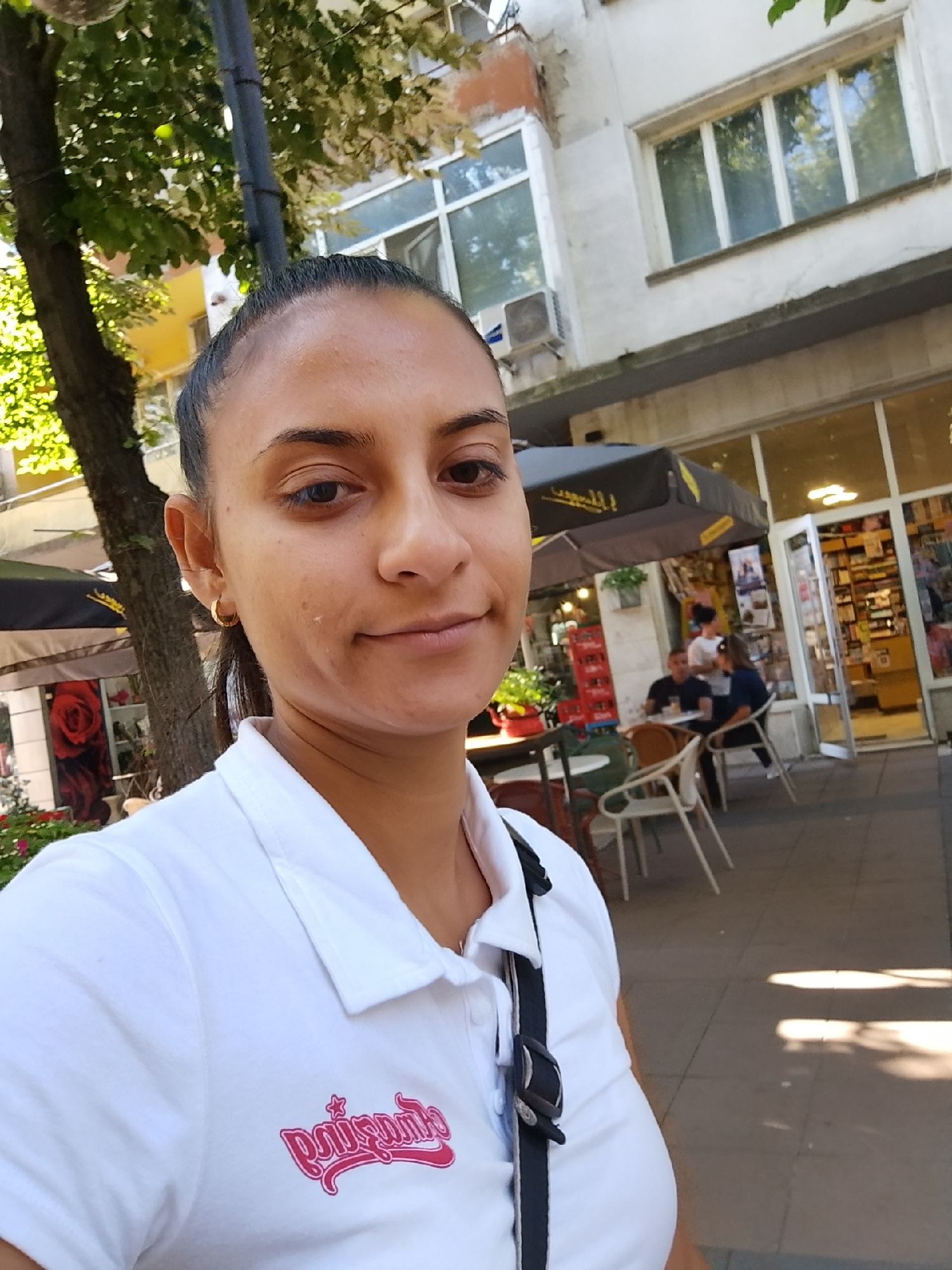 The image shows a woman in a white polo shirt with a pink logo on the left chest, taking a selfie outdoors. She has her hair pulled back in a ponytail and is wearing a gold hoop earring in her visible ear. Behind her, there is a building with white walls and windows, suggesting an urban setting. To the left of the woman, a tree with green leaves is visible, and further back, there are outdoor seating areas with umbrellas and tables, indicating a cafe or restaurant. People are seated at some of the tables, and a store with shelves of products can be seen through a glass door. This scene is located in Yambol, Bulgaria.
