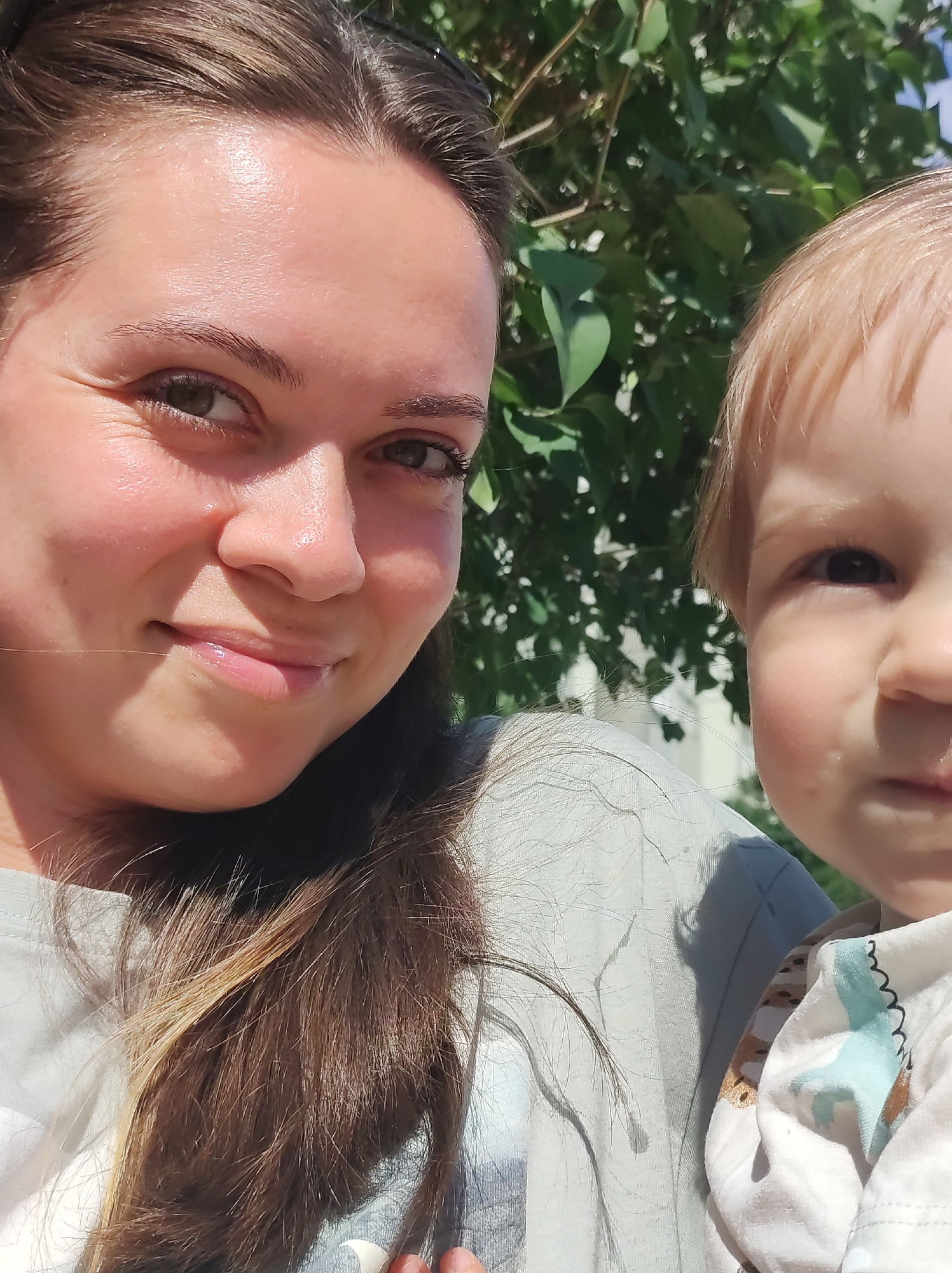 The image is a close-up, selfie-style photograph taken outdoors on a sunny day. On the left is a woman with medium-length brown hair, her face angled towards the camera with a slight smile. She is wearing a light grey t-shirt. To her right, a young child with blonde hair is partially visible, their face turned slightly away from the camera. The child is wearing a white garment with a pattern of animal figures. Behind them, lush green foliage and some sunlight filtering through suggest a natural, possibly park or garden, setting. There are no identifiable landmarks to suggest a specific location within the city of Chaikovsky, Russia.