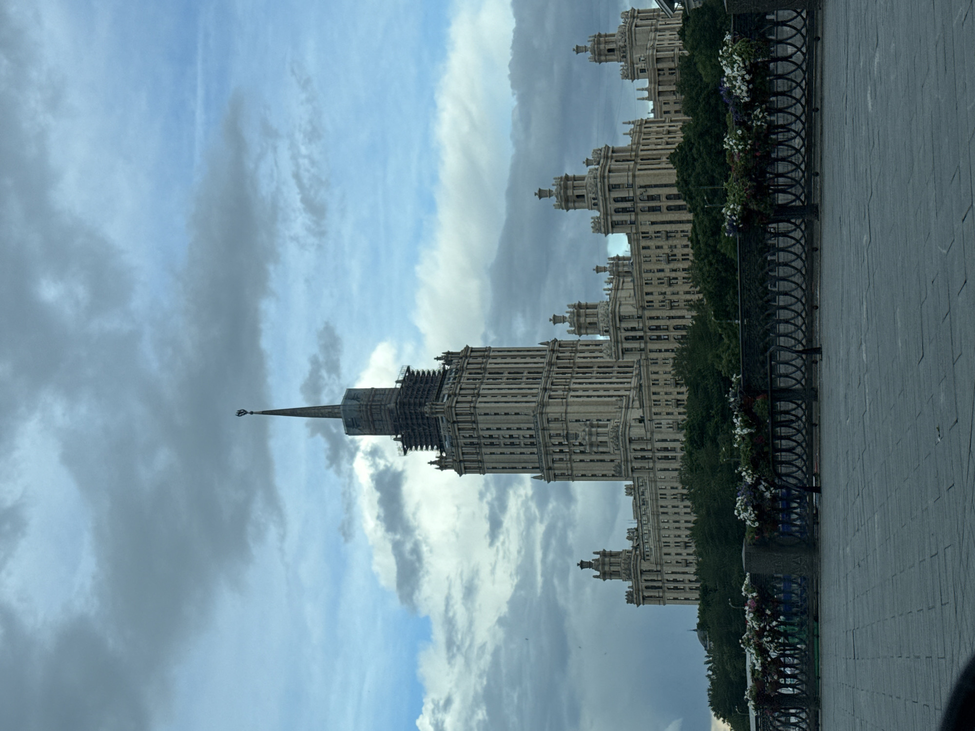The image depicts the Kotelnicheskaya Embankment Building in Moscow, Russia, captured from a low-angle perspective. The skyscraper rises prominently against a backdrop of a cloudy sky. A portion of the embankment walkway is visible in the foreground, including a decorative fence with flower-filled planters attached. The building’s architectural details are on display, including the multi-tiered structure, spires, and intricate façade. Some visible scaffolding near the top of the building indicates ongoing maintenance or construction. The overall setting suggests a cloudy day in Moscow.