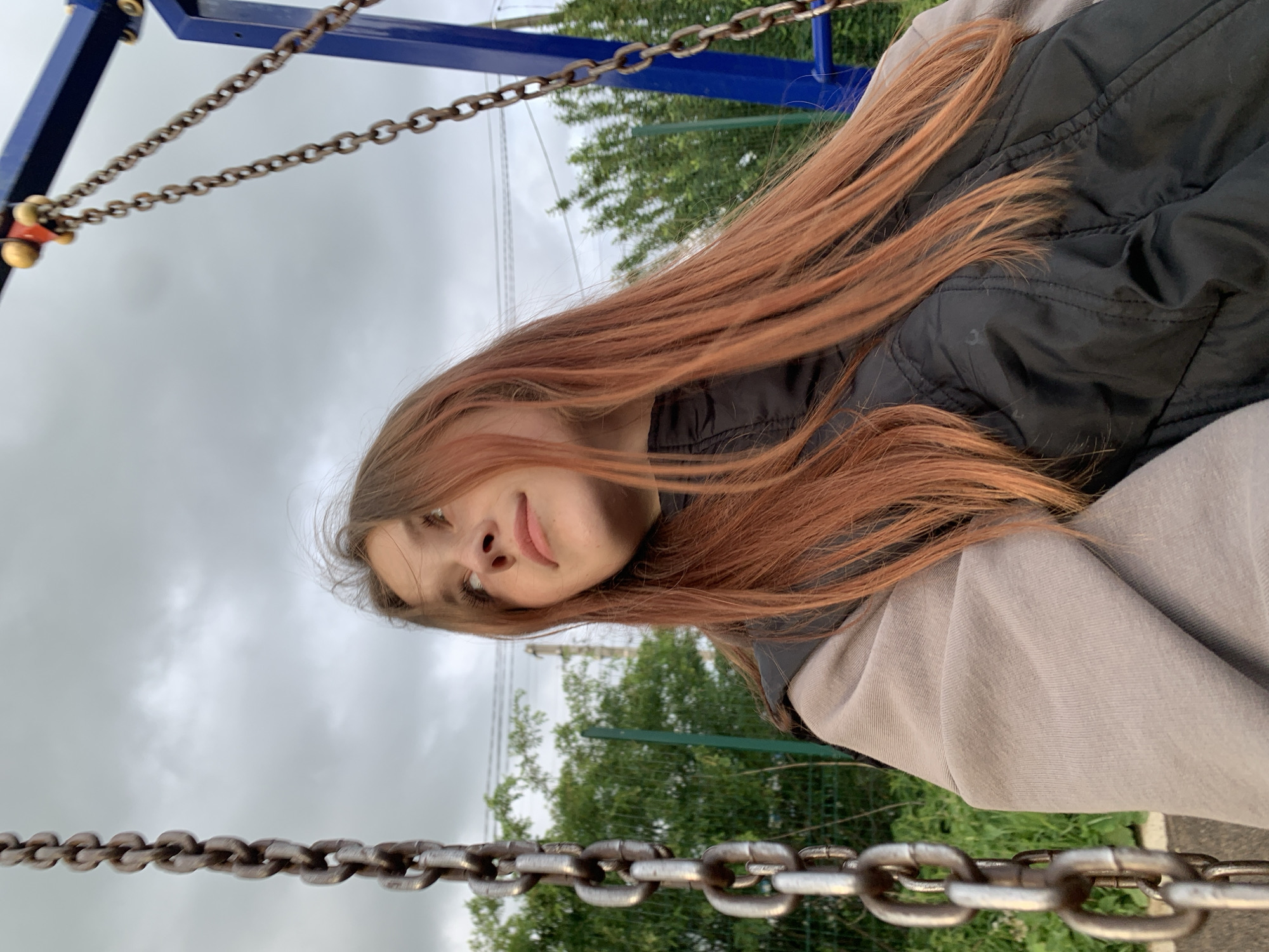 A young person, likely female, is depicted in an upward-facing, slightly angled shot, appearing to be lying or reclining on a swing and looking directly at the camera. She has long, flowing reddish-brown hair, which cascades downwards and across her black puffer jacket and a light grey hoodie or sweatshirt. A subtle, soft smile plays on her lips, conveying a relaxed or playful mood.
The perspective places the viewer below the subject. Thick metal chains from the swing set frame the bottom and top sections of the image, leading up to a blue metal swing frame visible in the upper background. The sky is overcast and cloudy, indicating daytime with diffused light. Green trees and other foliage are visible behind the swing structure, suggesting an outdoor setting like a park or playground. There is no visible text in the image. The scene takes place outdoors in Фоки, Russia.