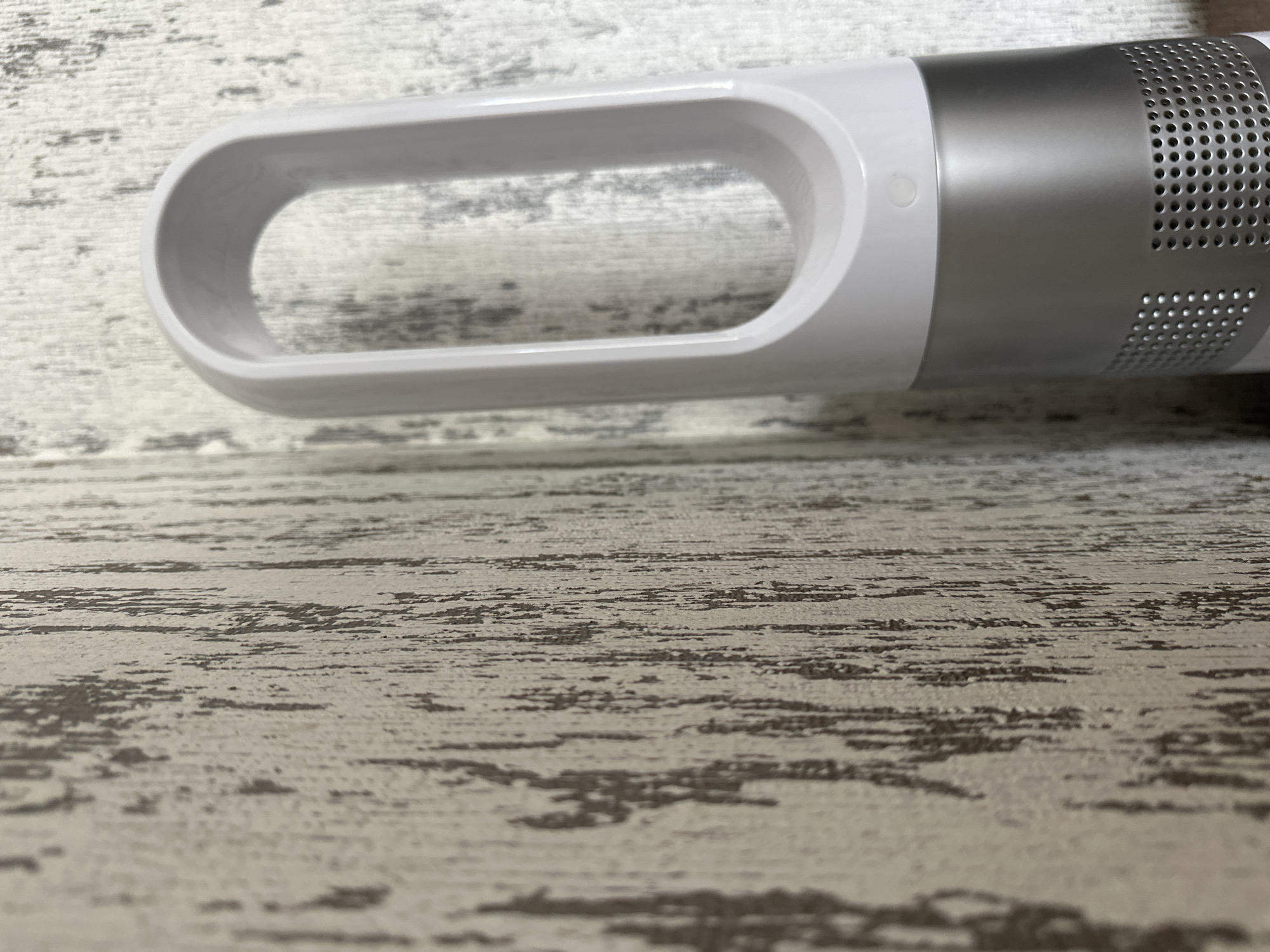 The image shows a close-up of a white and silver bladeless fan standing vertically next to a textured wall. The fan has a distinctive oval opening at the top and a cylindrical base with perforated holes, suggesting it is a modern design. The wall appears to have a distressed, vertical stripe pattern in shades of beige and grey. The lighting is soft, casting subtle shadows. The scene is indoors and static, with no people or animals visible. The provided context of Yessentuki, Russia, does not offer any visual cues to support or refute this location within the image itself.