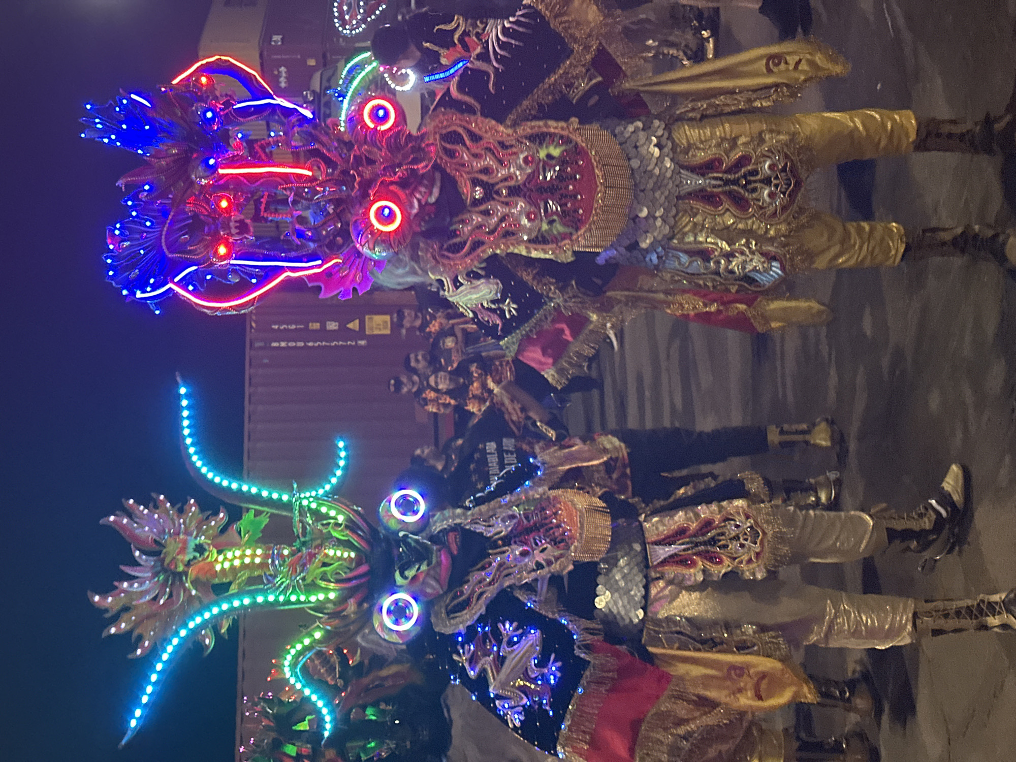 The image shows two costumed performers standing close together at night in Arica, Chile. The performers are wearing elaborate devil-like costumes, with large masks featuring illuminated eyes and horns decorated with LED lights. The costumes are heavily adorned with sequins, embroidery, and metallic fabrics in shades of gold, silver, and red. A group of people are visible in the background between the two performers. Behind them is an industrial-looking structure, possibly shipping containers. The background is dark, suggesting a nighttime setting.