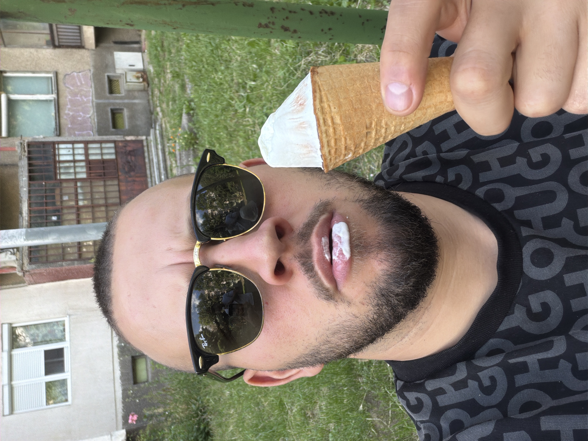 The image captures a close-up selfie of a male subject outdoors in Sofia, Bulgaria. The man has short dark hair and a dark beard, and is wearing black Ray-Ban Clubmaster-style sunglasses, reflecting an individual holding a phone. A smudge of white ice cream is visible on his lower lip. He holds a white ice cream in a waffle cone with his right hand. The subject is wearing a black t-shirt with a repeating grey "HUGO" letter pattern. The background features an urban setting with multiple older, multi-story buildings; one building to the left has a light-colored facade with a window and a closed external blind. Another building behind the subject shows rusted metal window grates and light-colored graffiti on a wall. The ground is covered in green grass with sparse vegetation. A vertical green metal pole is partially visible on the far right.