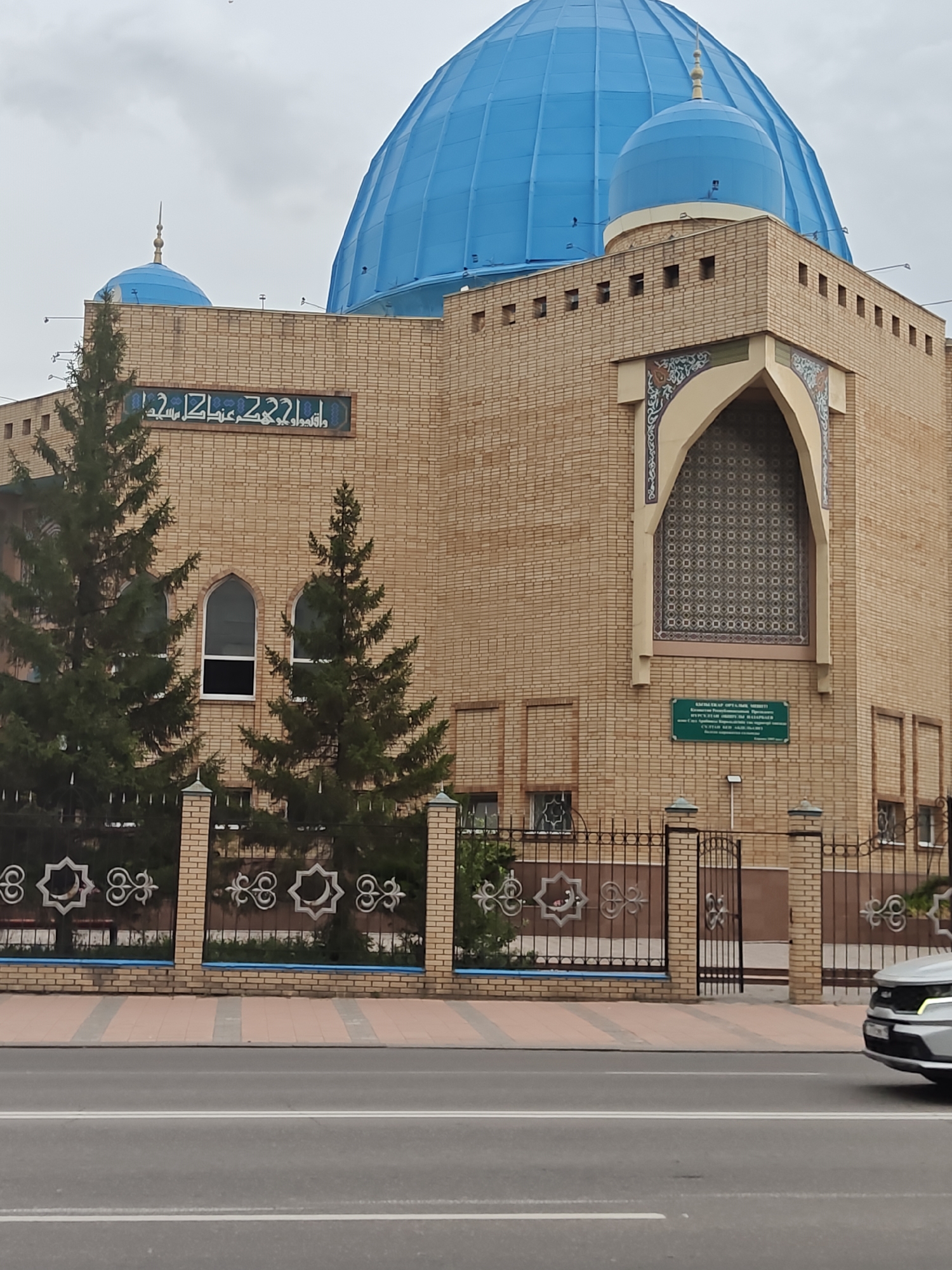 The image shows a modern mosque in the city of Petropavl, Kazakhstan. The mosque features a large blue dome and is constructed of light brown brick. Arabic script is visible above the arched entrance. Two tall evergreen trees stand in front of the mosque, partially obscuring the building. A decorative metal fence with a brick base separates the mosque grounds from the street. A silver car is partially visible on the right side of the image, driving on the asphalt road. The sky is overcast.