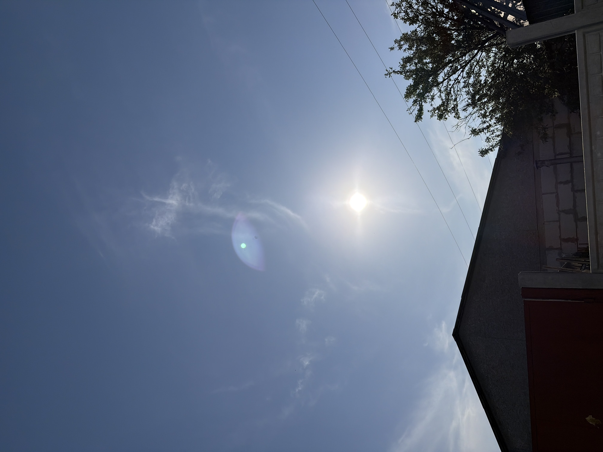 The image depicts a daytime scene in Chernihiv, Ukraine, under a clear blue sky with wispy clouds. The sun is prominently visible, casting a bright glare and lens flare. In the foreground, the dark, angled roofline of a building is silhouetted against the sky, with a red gate partially visible to the left. To the right, a dense green tree with visible branches extends upwards, and several power lines stretch horizontally across the frame. A portion of a brick wall structure is also present in the lower right section of the image.