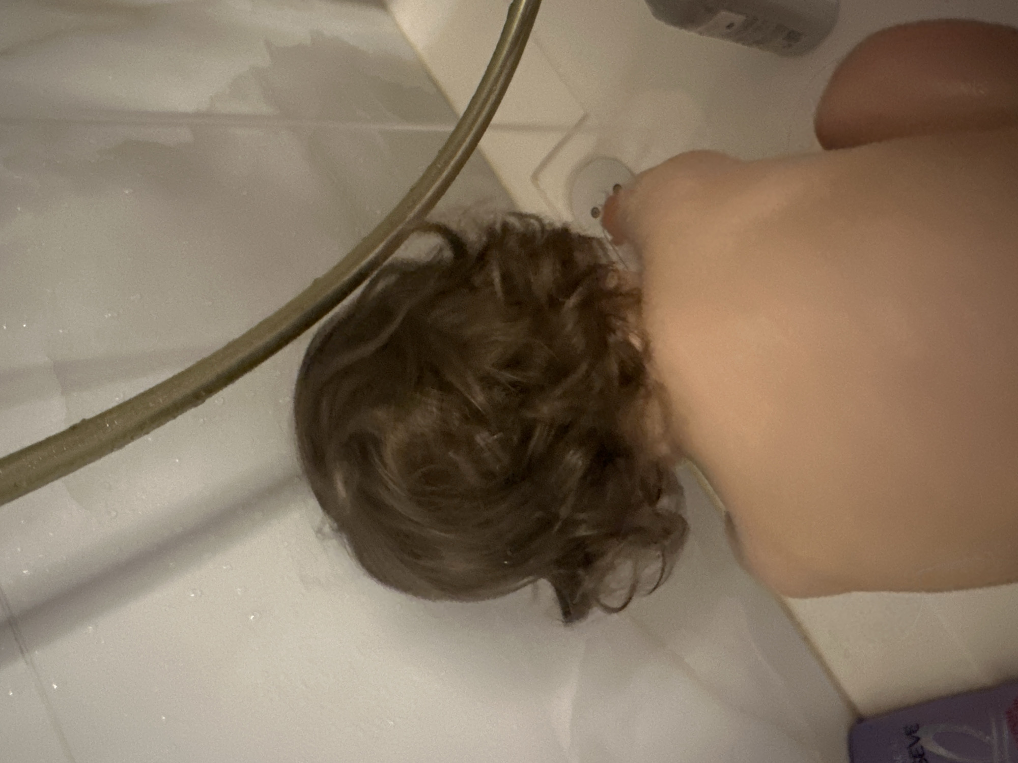A top-down view inside a shower stall or bathtub. A person's head with brown curly hair is visible under the shower head. A hand is pressing a button or dial on the wall. The scene takes place within a bathroom, possibly in an apartment or a private residence. The location of the image within the city of Khimki, Russia cannot be determined.