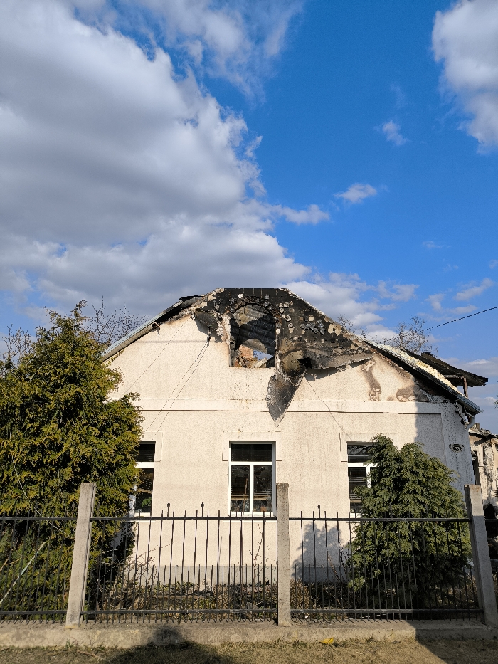 The image displays a two-story residential building located in Vinnytsia, Ukraine, exhibiting significant fire damage. The upper structure and gable roof are extensively charred and partially collapsed, with a large, irregular opening at the roof's peak exposing the damaged interior. Blackened debris and remnants of the roof structure are visible against the sky. The light-colored facade of the building shows soot and scorch marks primarily along the upper section, extending downwards from the roofline. Two intact windows with white frames are visible on the ground floor. A low black metal fence with concrete posts runs in the foreground, separating the building from the viewer. A dense green evergreen shrub obscures part of the building's left side, and a smaller shrub is present to the right. The sky overhead is blue with scattered white clouds. The scene is illuminated by daylight, with shadows cast by the fence posts.