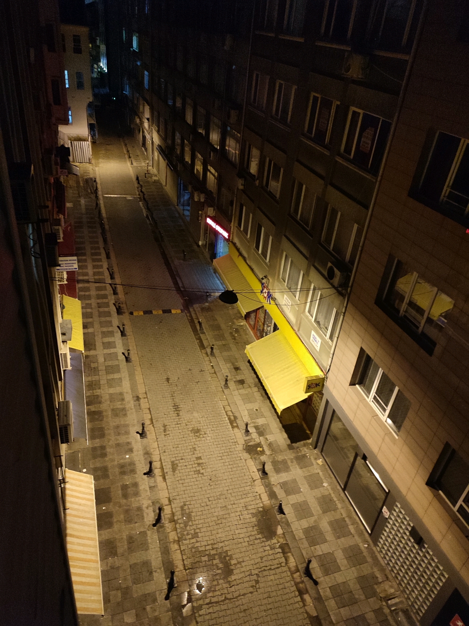 The image depicts a nighttime view looking down an alleyway in Kadıköy, Turkey. The alley is paved with cobblestones, some of which appear wet, indicating recent rain. Buildings line both sides of the alley. On the left, a building with several windows and an awning is visible. On the right, a larger building with multiple floors and numerous windows dominates the view. Some windows have signs or posters visible inside. Yellow awnings extend from some of the businesses on the ground floor of the right-hand building. Several bollards are placed along the edges of the sidewalk. The lighting in the alley is dim, with some artificial lights illuminating the street and the windows of the buildings. The scene is quiet and appears deserted.