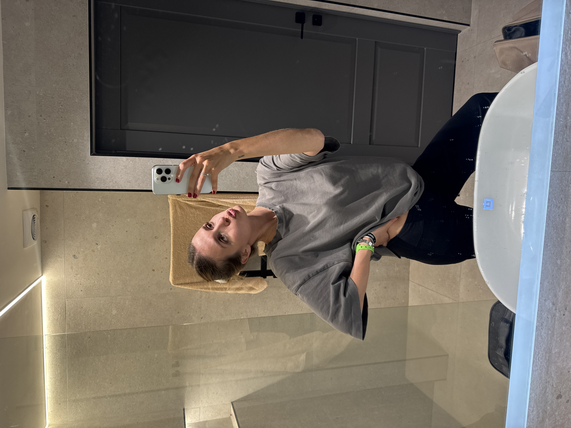 A woman is taking a selfie in a bathroom. The bathroom features a mirror, a door, and tiled walls. The location is most likely in Yerevan, Armenia, based on potential context cues not visible in the image. The woman is wearing a grey t-shirt and black leggings. She is holding a white smartphone with a visible camera setup. A towel is hanging on a rack behind her.