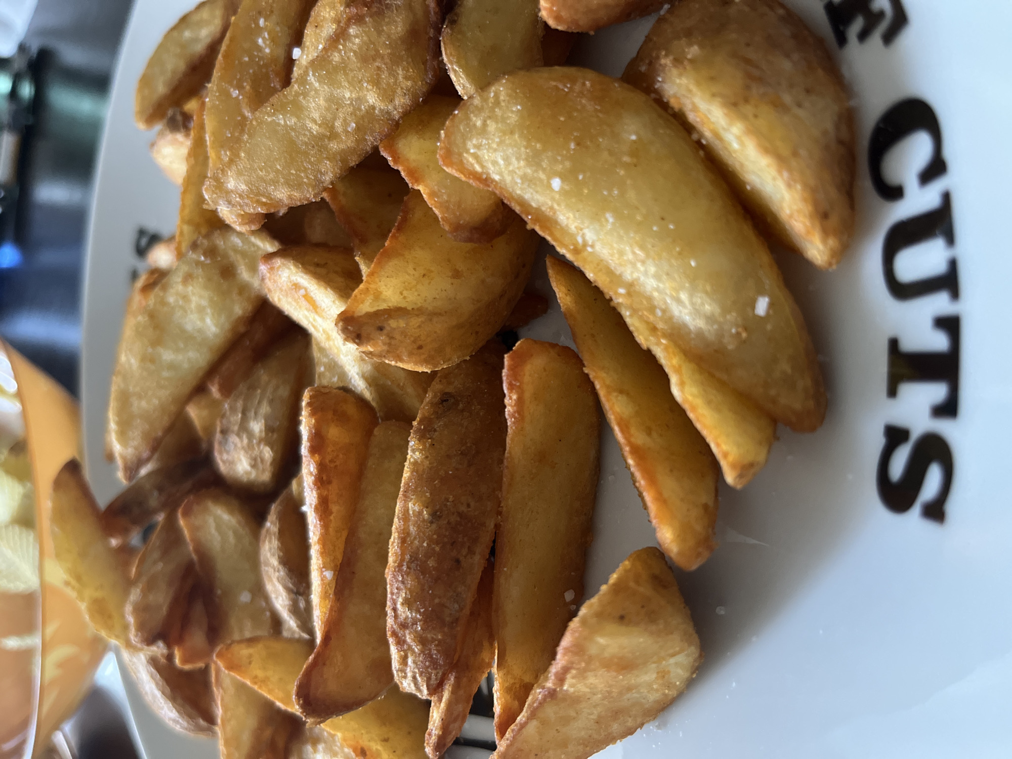 Within an indoor setting in the city of Rusanovo, Russia, a close-up perspective captures a white ceramic plate laden with multiple golden-brown potato wedges. These potato pieces exhibit irregular, rustic cuts, with some retaining their natural skin. Their surfaces appear crispy and textured, consistent with cooking processes such as frying or baking. Visible white, crystalline grains, identified as coarse salt, are unevenly distributed across the wedges. The plate's lower rim displays an inverted, black textual fragment, reading 'S CUTS'. The background is rendered out of focus, revealing a blurred orange-toned bowl on the left and an indistinct dark surface on the right. No individuals are present in the visible frame.