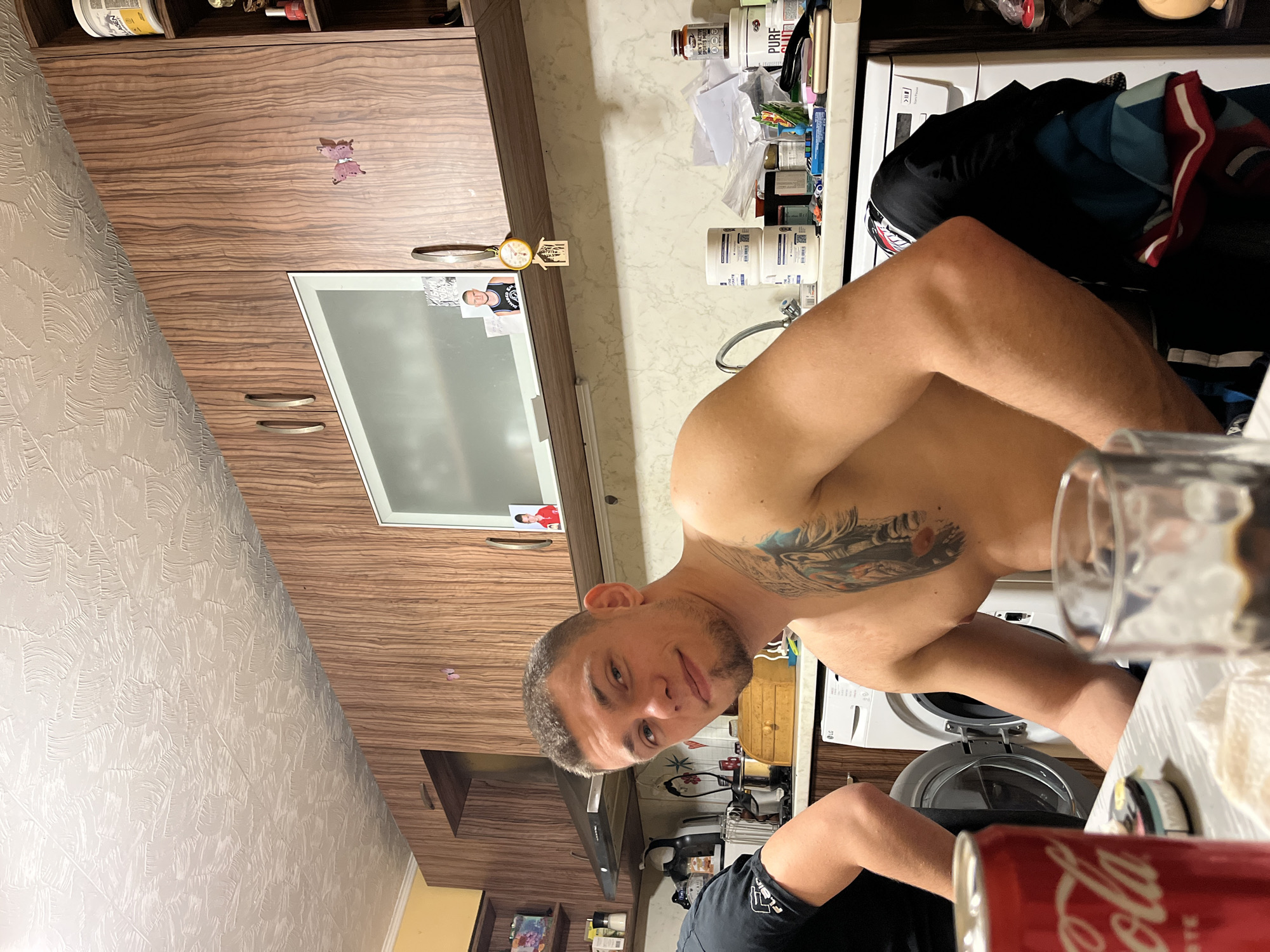 A man with a tattoo on his shoulder is standing in what appears to be a kitchen. The location is identified as Burgas, Bulgaria, based on unspecified contextual clues. The kitchen has wood-grain cabinets and a white countertop cluttered with various items, including medicine bottles and food products. A washing machine is visible. The man looks directly at the camera. A can of Coca-Cola is in the foreground.