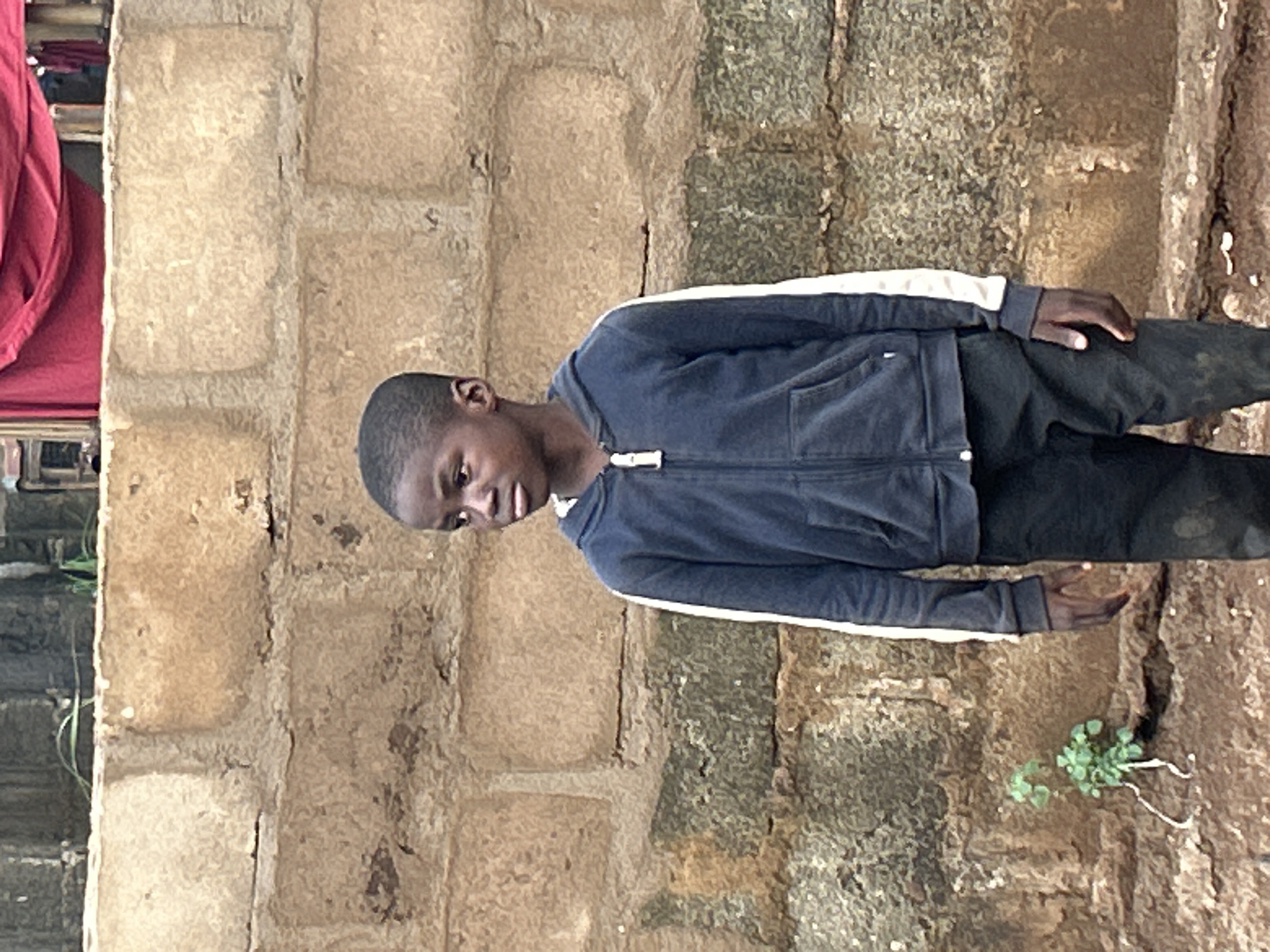 The image shows a young person, likely a boy, standing outdoors next to a rough-hewn, light brown brick or block wall. The original image is rotated 90 degrees clockwise, so viewing it correctly, the person is standing upright.
The subject is dark-skinned with short, dark hair. They are wearing a dark blue zip-up hoodie with white stripes running down the outer sides of the sleeves, and dark-colored trousers. The hoodie is zipped up. The person is looking downwards and slightly to their right. Their left hand is resting on their hip or side, and their right hand is slightly extended downwards towards the ground.
The background consists primarily of a textured wall made of rectangular, unevenly shaped blocks, possibly mud bricks or rough concrete, varying in shades of light brown and grey. To the person's right, the ground is visible, appearing earthen and uneven, with a small cluster of green plants growing from it. On the far left of the image, partially visible behind the wall, is a section of red fabric or material, which might be part of furniture or another structure, along with some indistinct dark elements.
The lighting suggests it is daytime, with no strong shadows indicating harsh sunlight. The person's expression is relatively neutral or perhaps slightly pensive as they look down. There is no visible text in the image.