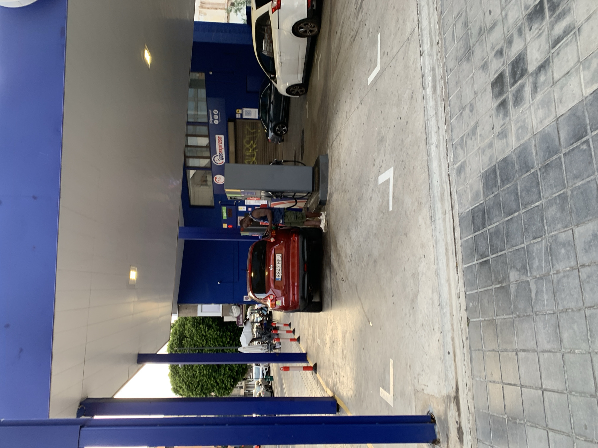The image depicts a scene at a gas station in Valencia, Spain. In the foreground, a red Nissan Juke with license plate "8847 HFJ" is parked, its rear facing the camera. To its right, a Black man in a blue t-shirt and olive-green shorts stands next to a gas pump, holding a mobile phone. A white minivan is parked behind the red car. Further back, a dark-colored sedan is also at a pump. The gas station has blue architectural elements, including a prominent canopy. White directional arrows are painted on the ground. In the background, trees and buildings are visible. The ground surface transitions from concrete to tiled pavement.