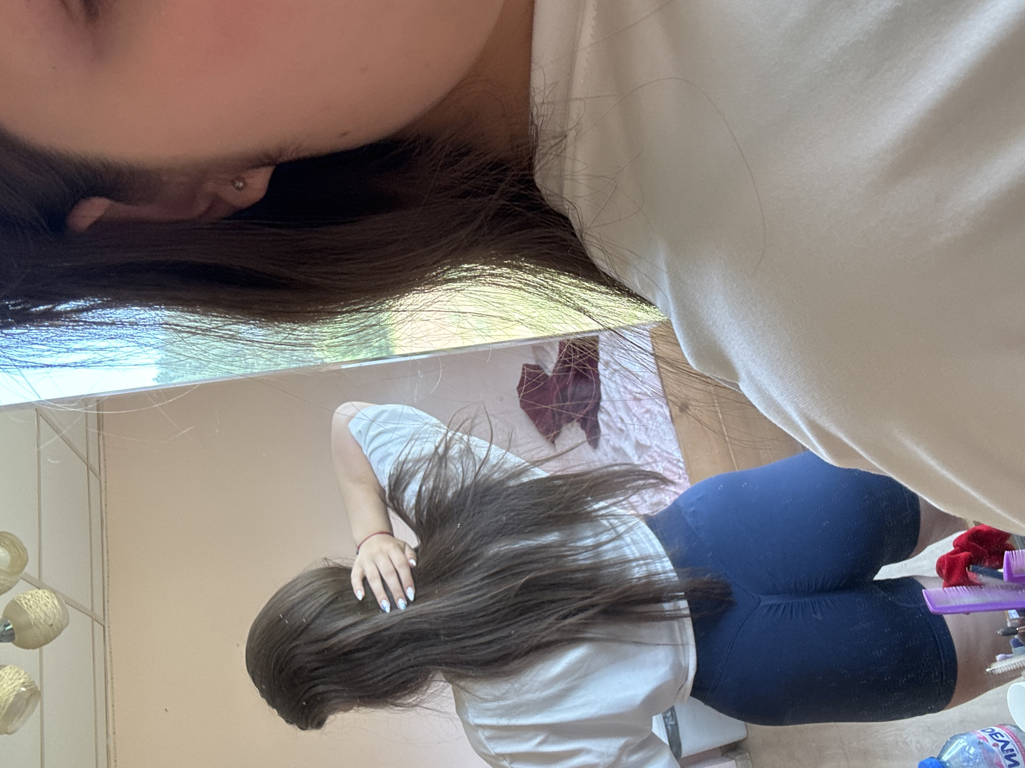 A young woman with long, dark brown hair is standing in front of a mirror, taking a selfie. She is wearing a white t-shirt and dark blue athletic shorts. Her hair is styled in a way that she is holding it up with one hand, revealing her back. Her nails are painted with a French manicure. In the reflection of the mirror, we can see a room with a pink bedspread, a burgundy blanket, and a light-colored ceiling with three decorative lamps. The woman is also wearing a small stud earring in her visible ear. A bottle of water and some hair accessories are visible on a surface in the foreground. The overall lighting suggests daytime.