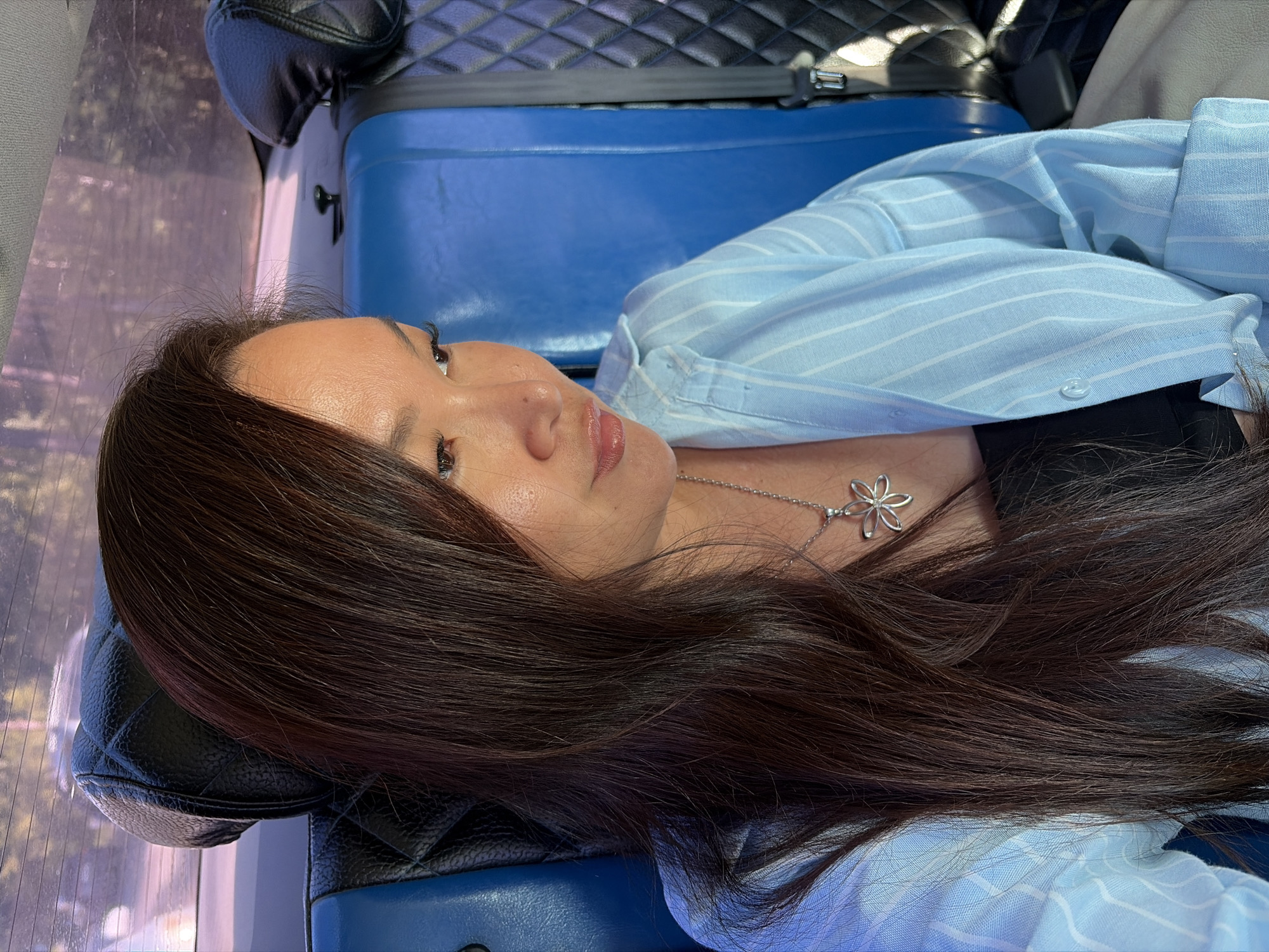 The image shows a woman with long, dark brown hair, wearing a light blue and white striped button-down shirt over a black top. She is also wearing a silver pendant necklace with a flower-shaped charm. She is sitting in the back seat of a vehicle, with blue seats and quilted black headrests visible. Sunlight is streaming in from the side, casting shadows and highlighting her face. The background appears to be a blurred view of trees and sky through the rear window of the vehicle. The scene is consistent with being inside a car or bus on a sunny day, and the woman is looking down and to her right. There is no specific indication of the location being in Chaikovsky, Russia, beyond the prompt's instruction.