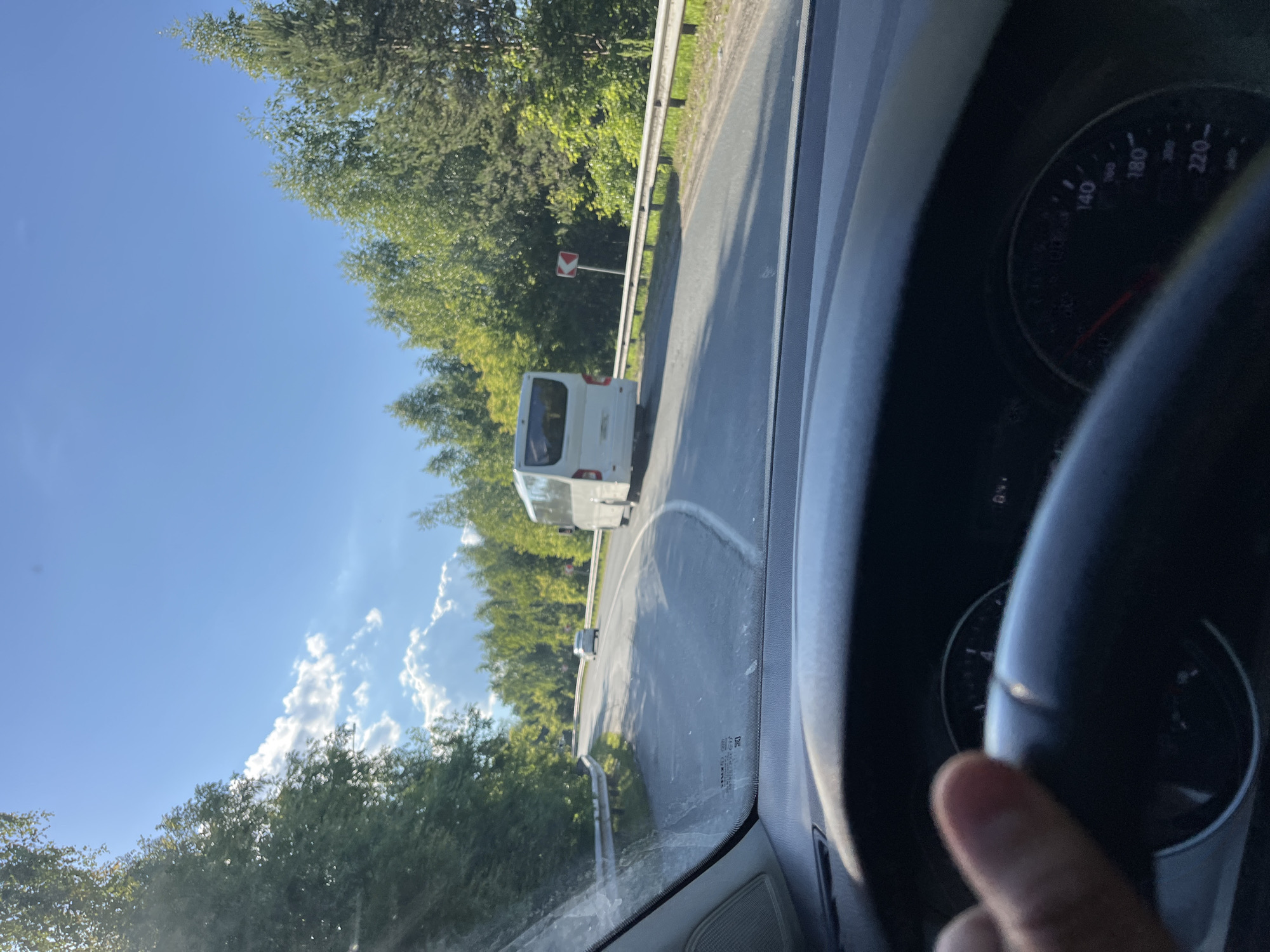 The image is a first-person perspective from inside a vehicle, likely a car, looking out through the windshield. The vehicle is on a paved road that curves to the left. In the foreground, the dashboard and part of the steering wheel are visible. A hand is resting on the steering wheel. The road ahead is flanked by dense green trees on both sides.
A white bus is driving ahead on the road, and a smaller white car is further in the distance. There is a speed limit sign with a red border and an arrow indicating a curve to the right, positioned on the right side of the road. The sky is blue with scattered white clouds. The lighting suggests it is daytime, possibly late afternoon due to the angle of the sun.
The image may have been taken on a road near the city of Vasilyevo, Russia, given the context. The overall environment is rural or suburban, with a focus on the road and the surrounding natural landscape.