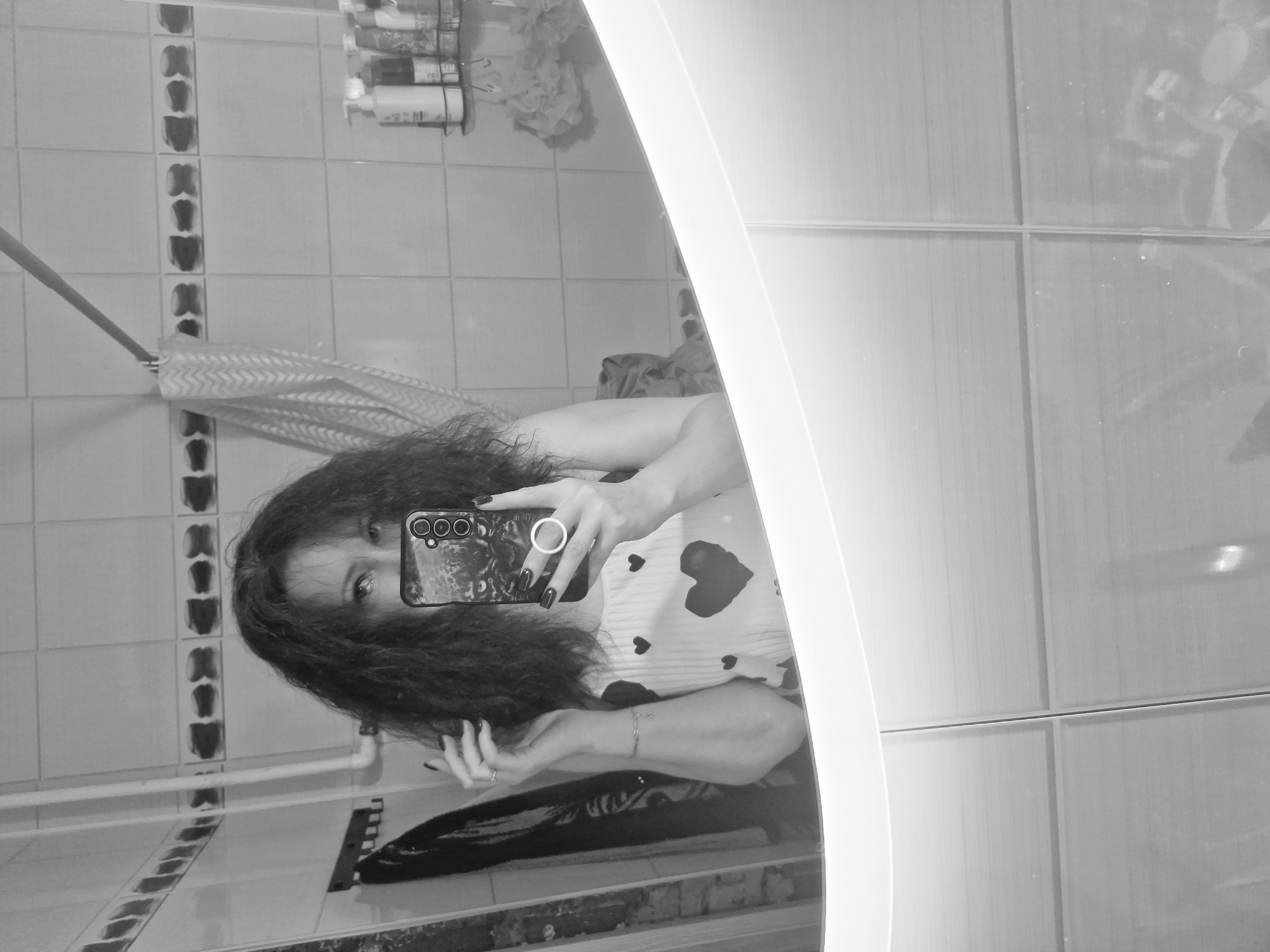 A person stands in a bathroom in Марковский, Russia, holding a smartphone and taking a mirror selfie. The bathroom has tiled walls and a curved, illuminated mirror. A shower rod, a towel, and shower products are visible in the background. The person is wearing a shirt with heart patterns.