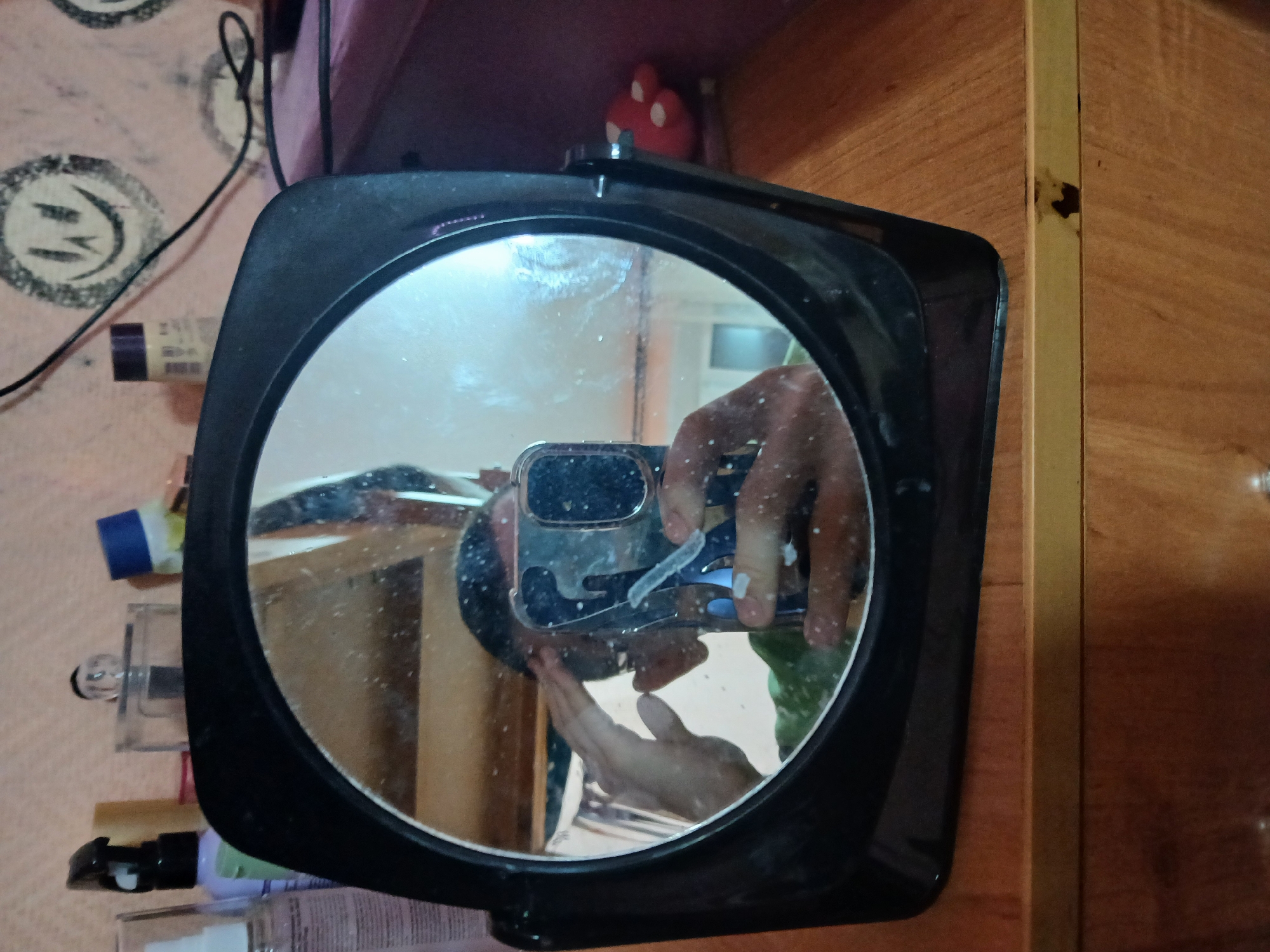 A person is taking a picture of themselves in a circular mirror with a black frame. The reflection shows a hand holding a smartphone with a clear case. In the background, various cosmetic products are visible on a surface covered with a patterned cloth. To the right of the mirror, a wooden cabinet or wall is present. Above the mirror, a small red object is positioned. The location is determined to be in the city of Chaikovsky, Russia.