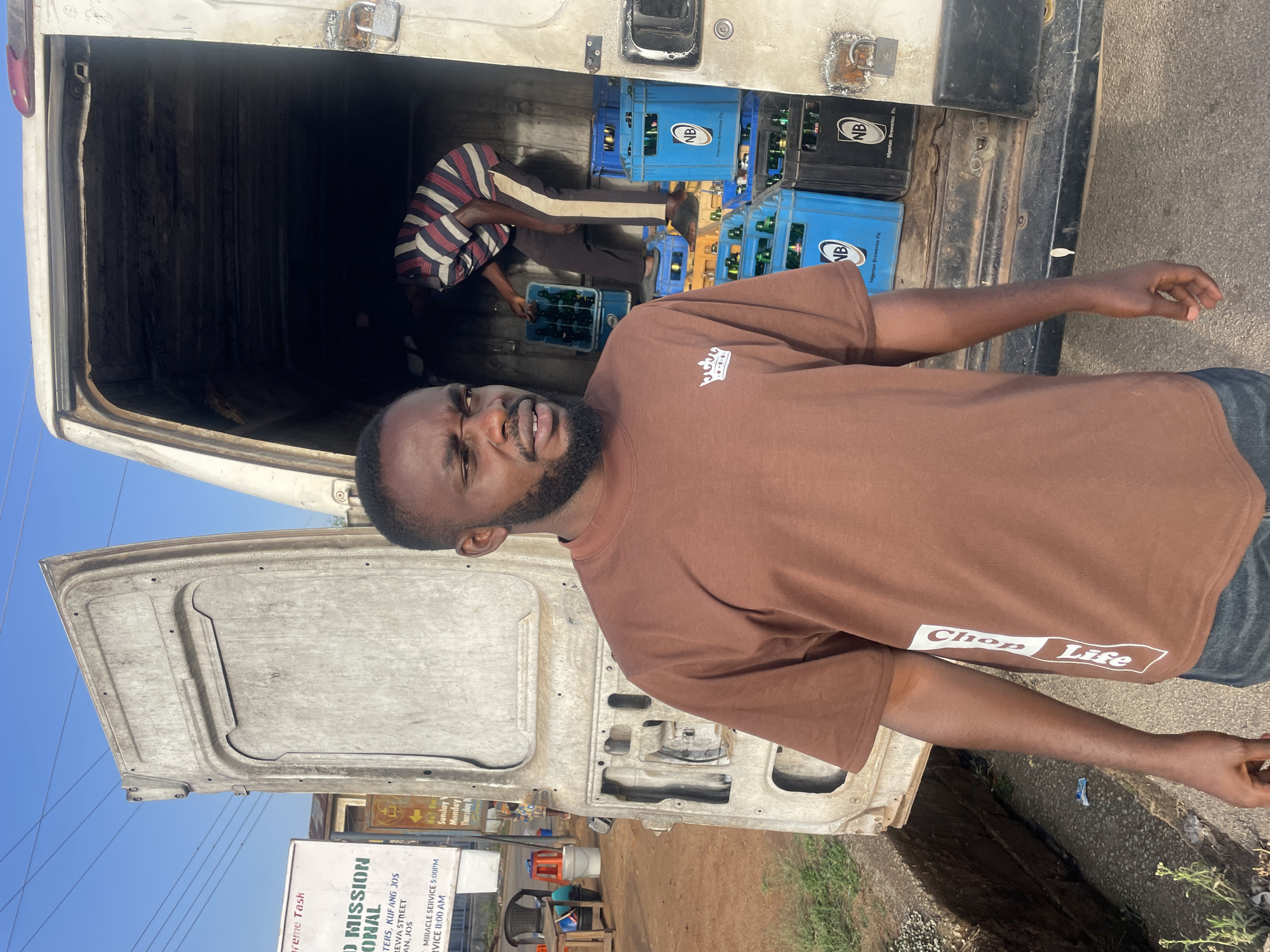 A man in a brown "Chop Life" T-shirt stands next to an open delivery truck in Jos, Nigeria. Inside the truck, a person in a striped shirt is handling crates of "NB" Nigerian Breweries beer bottles. A sign for "Word Mission International" on Kurewa Street, Jos, is visible.