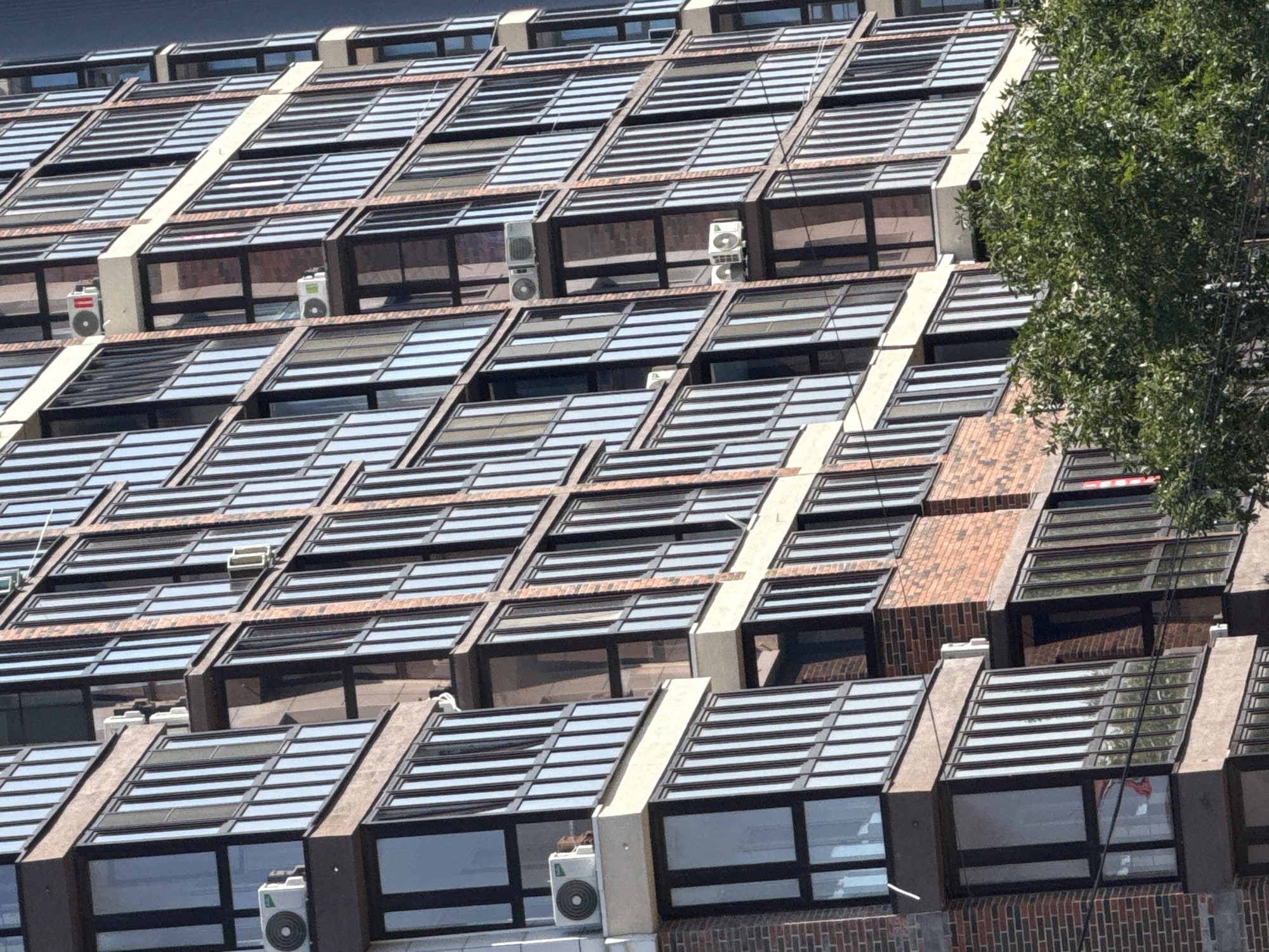 The image shows a close-up, angled view of a modern, multi-story residential building, likely an apartment complex, in Anapa, Russia. The building features a facade of dark brick and numerous glass-enclosed balconies or sunrooms with dark frames. Several white air conditioning units are mounted externally on the building, some on balconies and others directly on the wall. The sunlight creates strong reflections on the glass surfaces, obscuring the interior of the apartments. A lush green tree partially obstructs the lower portion of the building. The scene appears to be captured during daylight hours.