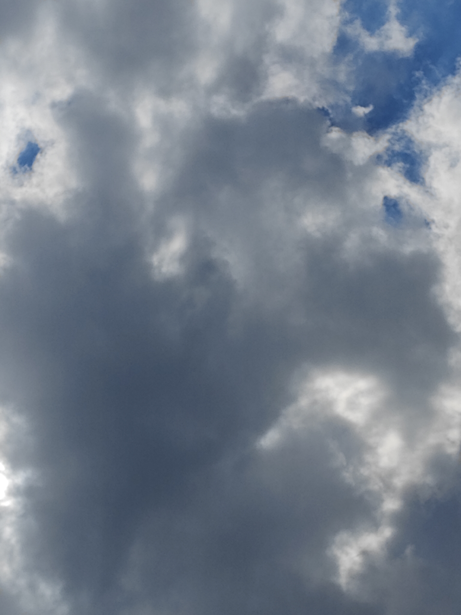This is a close-up, outdoor, eye-level shot of the sky filled with clouds. The clouds are a mix of dark gray and white, with patches of bright blue sky visible. The lighting suggests it might be daytime, possibly a partly cloudy or stormy day. There are no people, objects, or specific landmarks visible, making it impossible to determine a location within any city. The image focuses solely on the atmospheric conditions.
