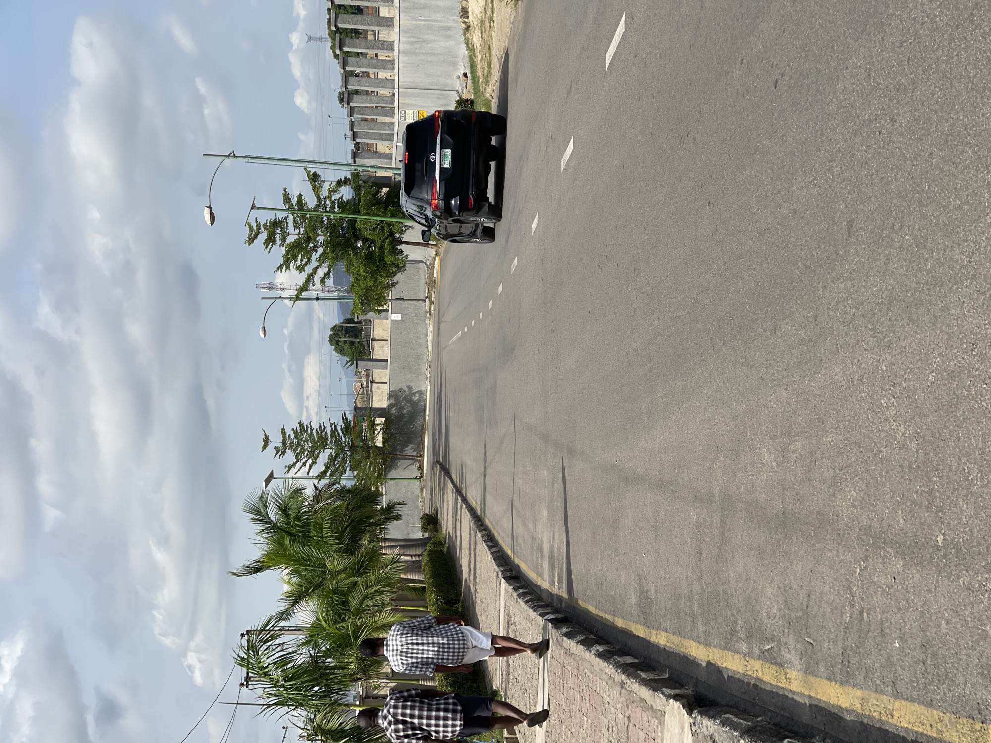 The image depicts a street scene in Gosa, Nigeria. A black Toyota SUV is driving on the road, heading away from the viewer. Two people are walking on the sidewalk beside the road. Palm trees and other trees line the street. Streetlights are also present. Buildings can be seen in the background. The sky is cloudy.