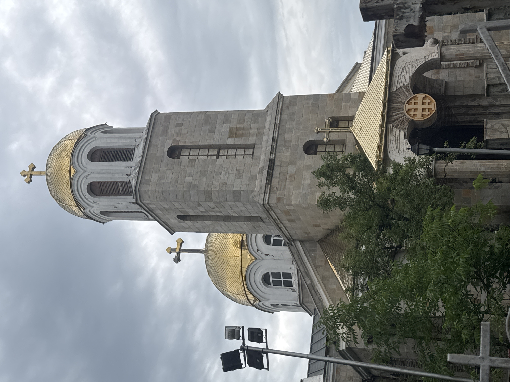 This media file captures an exterior view of a stone church with golden domes. The architecture is characterized by its use of stone blocks and arched windows. A prominent feature is the golden dome, topped with a golden cross, which reflects the overcast sky. Adjacent to the dome is a tall, slender bell tower, also constructed from stone.
The scene is set against a cloudy, grey sky, suggesting it might be daytime but with overcast weather. The lighting is diffused, creating a somber or perhaps peaceful atmosphere. There are some green trees visible in the foreground, adding a touch of nature to the predominantly architectural subject. The image also shows some modern lighting fixtures on poles, suggesting the presence of infrastructure around the church.
Notable details include the intricate stonework and the metallic sheen of the golden domes and crosses. The overall impression is one of solemnity and grandeur, typical of religious architecture. No people are visible in the frame, and no text is discernible. The image appears to be taken from a slightly upward angle, emphasizing the height and scale of the church.