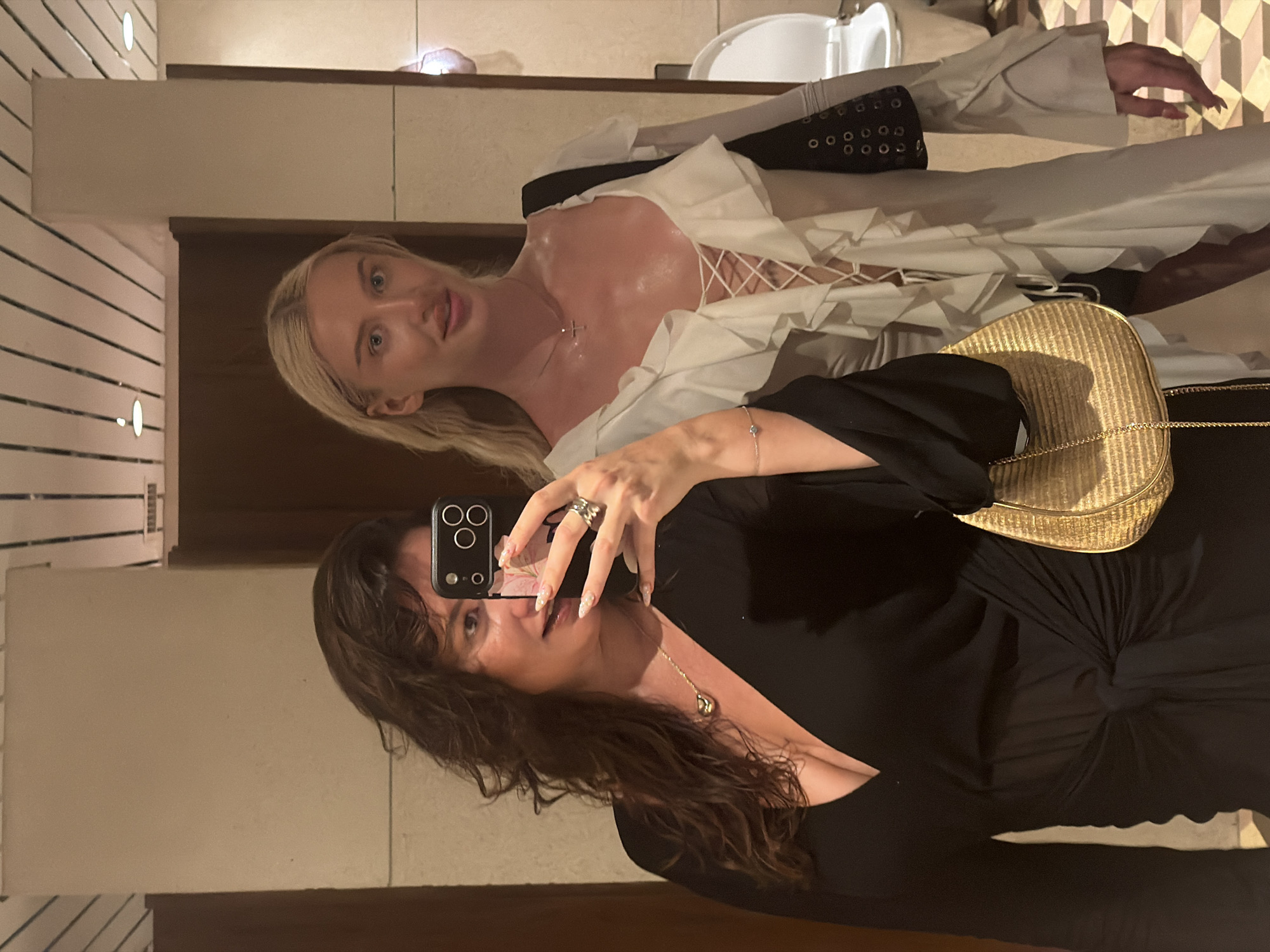 In Kuta Selatan, Indonesia, two women are seen taking a mirror selfie in what appears to be a well-lit, tiled restroom. The woman on the left, with dark, wavy hair, is wearing a black dress with a deep V-neck and a knotted detail at the waist. She is holding a smartphone in her right hand, capturing the reflection. A gold-colored, woven handbag with a chain strap is visible on her left side. She is wearing jewelry, including a pendant necklace and rings.
The woman on the right, with blonde hair, is wearing a white ruffled dress with a lace-up front and voluminous sleeves. A silver necklace with a cross pendant is visible on her neck. She is holding a black bag with silver grommets over her right shoulder. In the background, a white toilet and a tiled floor are visible. The walls are paneled with what looks like a light-colored material. The overall lighting suggests an indoor setting.