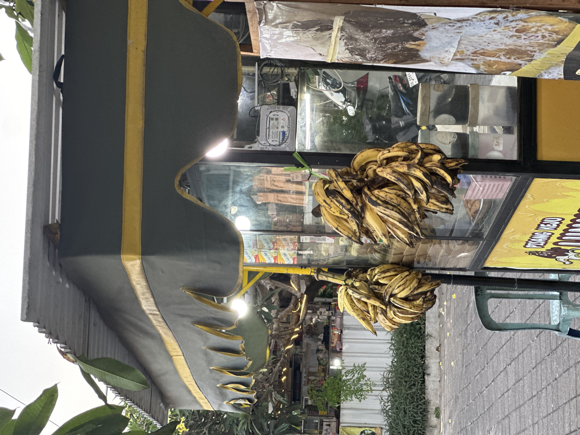 A street food kiosk selling bananas is located in Kuta Selatan, Indonesia. The kiosk has a green and yellow awning, and bunches of bananas hang from the side. Inside the kiosk, there are shelves stocked with goods, and a glass display case shows various items. A blue plastic chair sits in front of the kiosk. The ground is paved with gray bricks. In the background, there are trees and other buildings. A motorcycle is parked behind the kiosk.