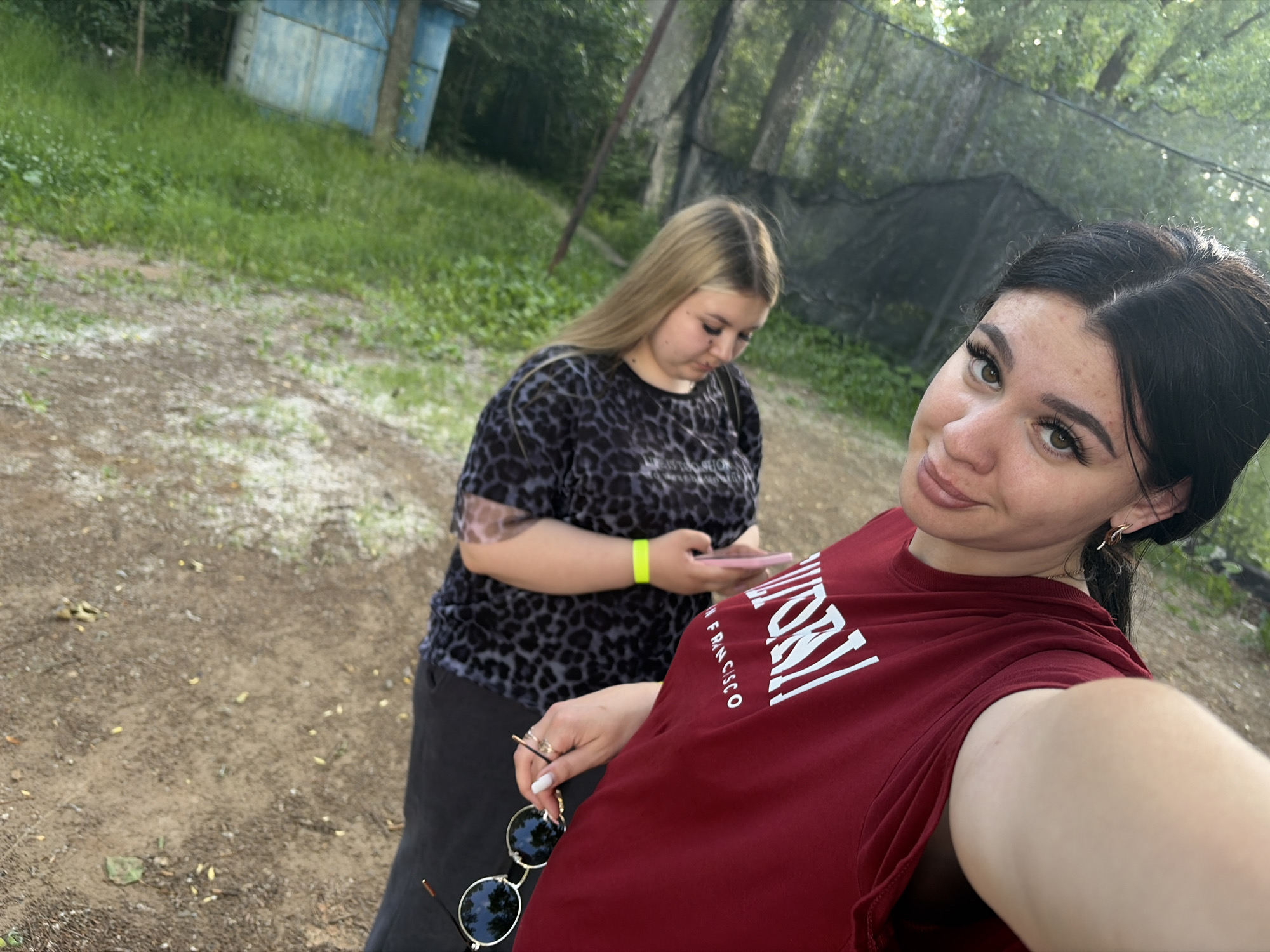 Two young women are captured in a selfie-style shot outdoors. The woman in the foreground, with dark hair and makeup, is wearing a maroon t-shirt with white text that appears to read "UNIVERSITY OF SAN FRANCISCO". She is smiling and holding the camera with her right arm. Her left hand is visible, holding a pair of sunglasses and a ring. The second woman, with blonde hair and wearing a black and grey leopard-print shirt and dark pants, is standing behind and to the left of the first woman. She is looking down at her phone, which she is holding in her right hand, and has a bright yellow wristband on her left arm.
The setting appears to be a wooded area with a dirt path and some grass. In the background, there is a blue structure that resembles a shed or small building, and a dark netting structure that could be part of a sports field or enclosure. The overall impression is of a casual outdoor gathering or outing. The lighting suggests it is daytime, possibly overcast or in the late afternoon given the diffused light. The presence of the netting and the overall rustic environment might suggest an activity like paintball or an adventure park. The women appear relaxed and are engaged in their own activities, with the foreground woman sharing a pleasant moment with the camera. There are no strong indicators of extreme weather conditions.