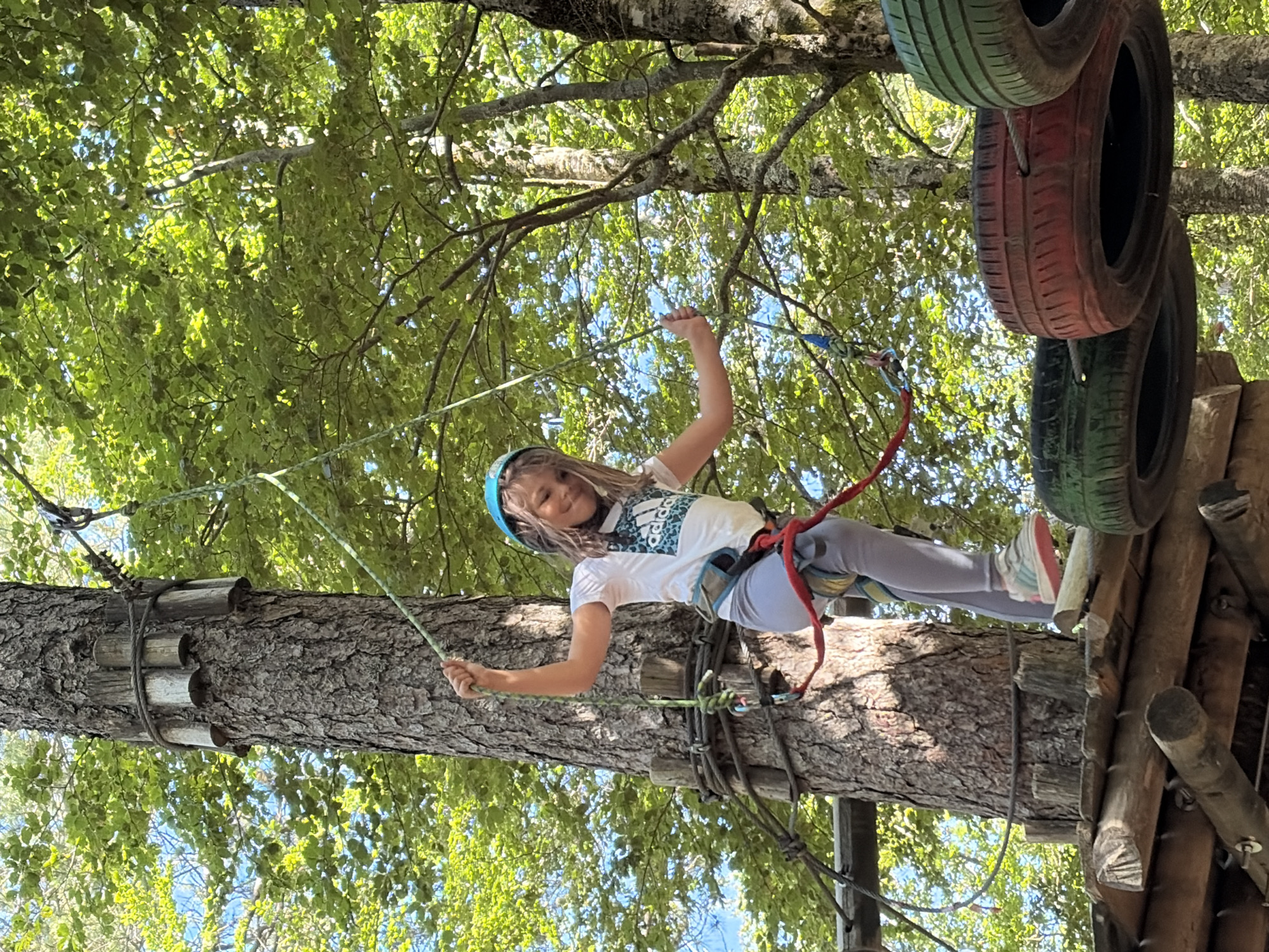 A young girl, estimated to be between 8 and 12 years old, actively participates in an aerial adventure course within a forest located in Mali Bostur, Montenegro. She is positioned on a wooden platform attached to a large tree trunk, which has dark, textured bark. The girl wears a blue protective helmet, a white short-sleeved t-shirt with a teal and black Adidas logo on the chest, light grey leggings, and light-colored athletic shoes with pink accents. A red and blue safety harness is visible around her waist and legs. She grasps a thick green safety rope with both hands, extending her arms upwards. Her facial expression indicates a smile. The tree trunk is outfitted with horizontal wooden logs secured by ropes, forming part of the course infrastructure. To the right of the girl, a series of recycled tires, painted red, green, and black, are suspended by cables, forming a subsequent obstacle. The background is dominated by lush green foliage of numerous trees, indicating a dense forest environment, with intermittent patches of blue sky visible through the canopy. Sunlight illuminates parts of the scene, creating bright highlights on the leaves and the girl's clothing.