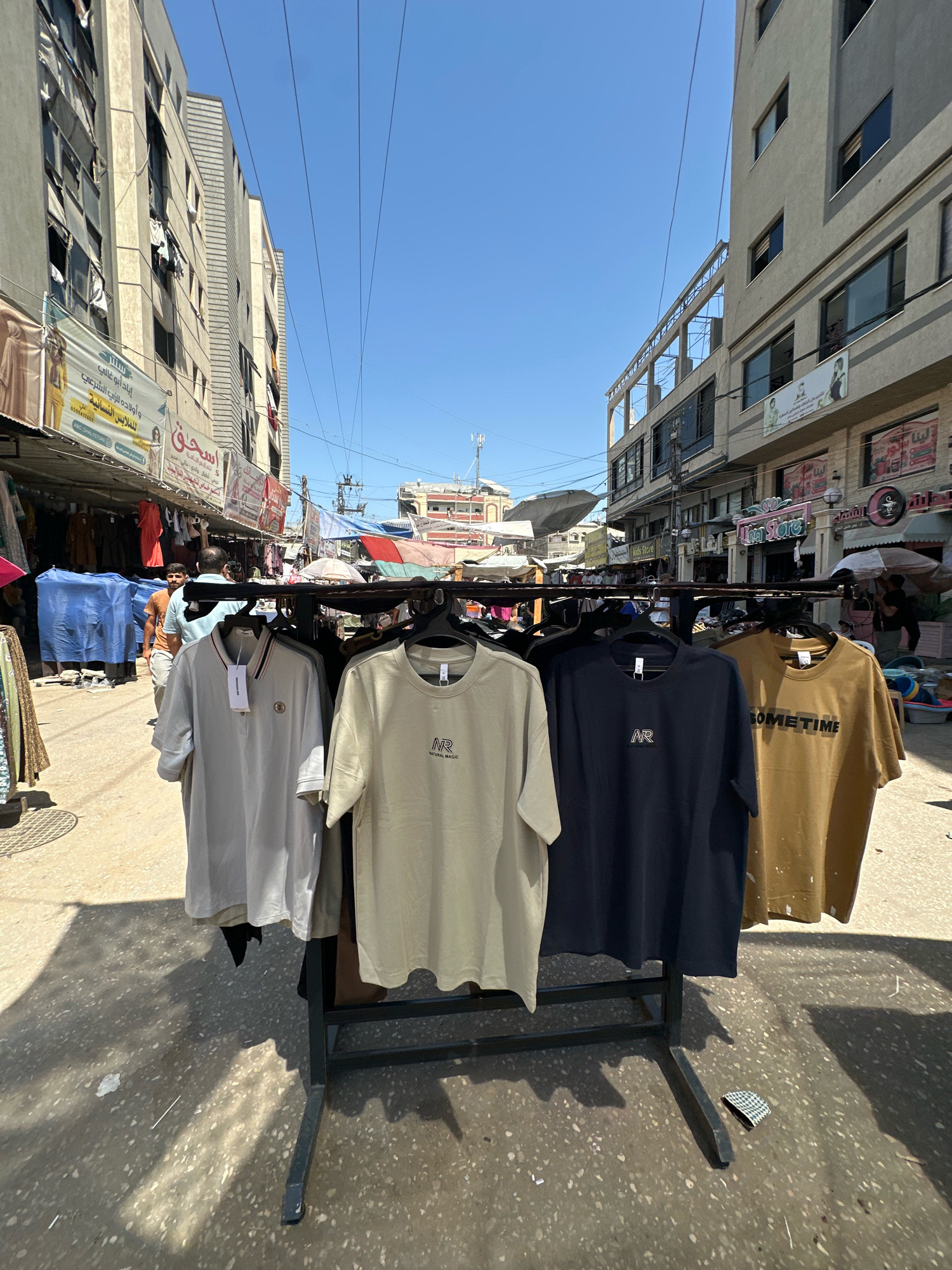 The image depicts an outdoor market scene in Nuseirat, Palestinian Territories. The foreground features a black metal rack displaying four t-shirts: a light grey polo shirt with a striped collar, a pale green t-shirt with a small "NR" logo, a navy blue t-shirt with the same "NR" logo, and a brown t-shirt with the text "SOMETIME" in black lettering. The t-shirts are hung on hangers. The ground is a textured, speckled concrete surface with shadows cast by the t-shirts and overhead structures.
In the background, a bustling street market is visible. Buildings with multiple floors line both sides of the street, some appearing weathered with visible damage to windows and facades. Banners and signs with Arabic writing are displayed on the buildings. Stalls with various goods, including clothing and other merchandise, are set up along the street. Several people are seen walking through the market. Blue tarps cover some items. Overhead, power lines crisscross the sky, which is clear and blue. The overall impression is of a busy, everyday market in an urban environment.