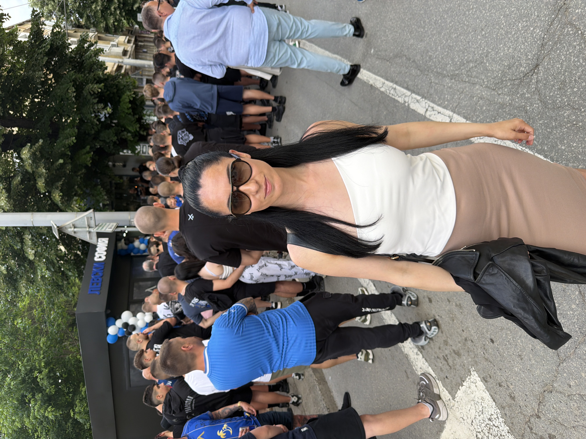 A woman in sunglasses, wearing a white tank top and tan skirt, stands facing the camera on a street in Pleven, Bulgaria. She is holding a black leather jacket. Behind her is a crowd of people gathered near a building labeled "ЛЕВСКИ СОФИЯ" (Levski Sofia). The building is decorated with white and blue balloons. Trees are visible in the background. A street is visible in the front.
