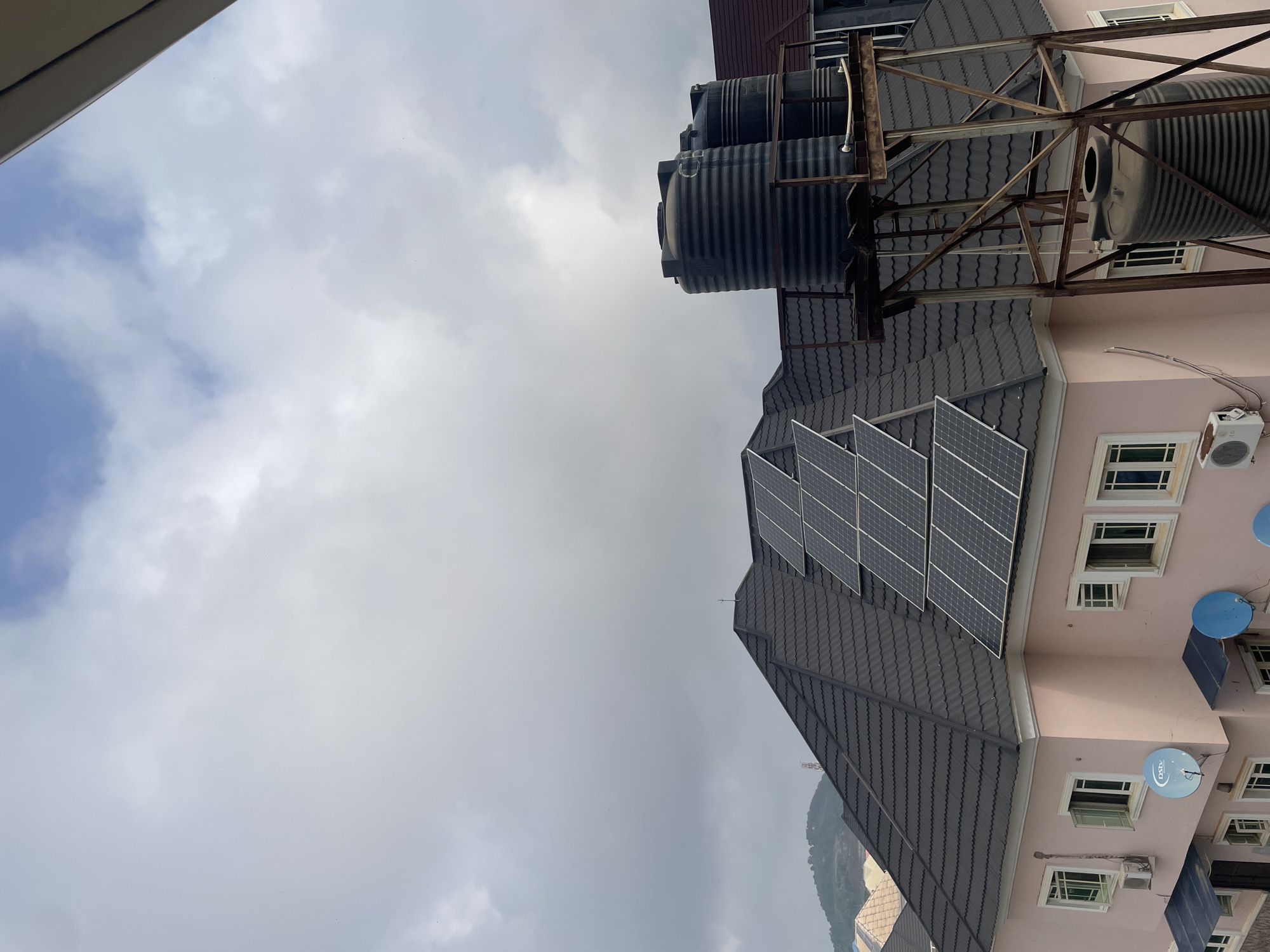 This image captures a scene from the city of Abuja, Nigeria, focusing on residential rooftops. The foreground features a pink, multi-story building with a dark, tiled roof. Several solar panels are strategically placed on this roof. Adjacent to the building, a rusty metal structure supports two large, black water tanks stacked vertically, with a third tank positioned lower on the structure. The building has multiple windows, some open, and air conditioning units are visible. Two blue satellite dishes are mounted on the side of the building. In the background, more residential buildings with tiled roofs are visible, along with rolling hills under a cloudy sky. The overall impression is one of modern residential infrastructure, likely in a suburban or developed urban area within Abuja.