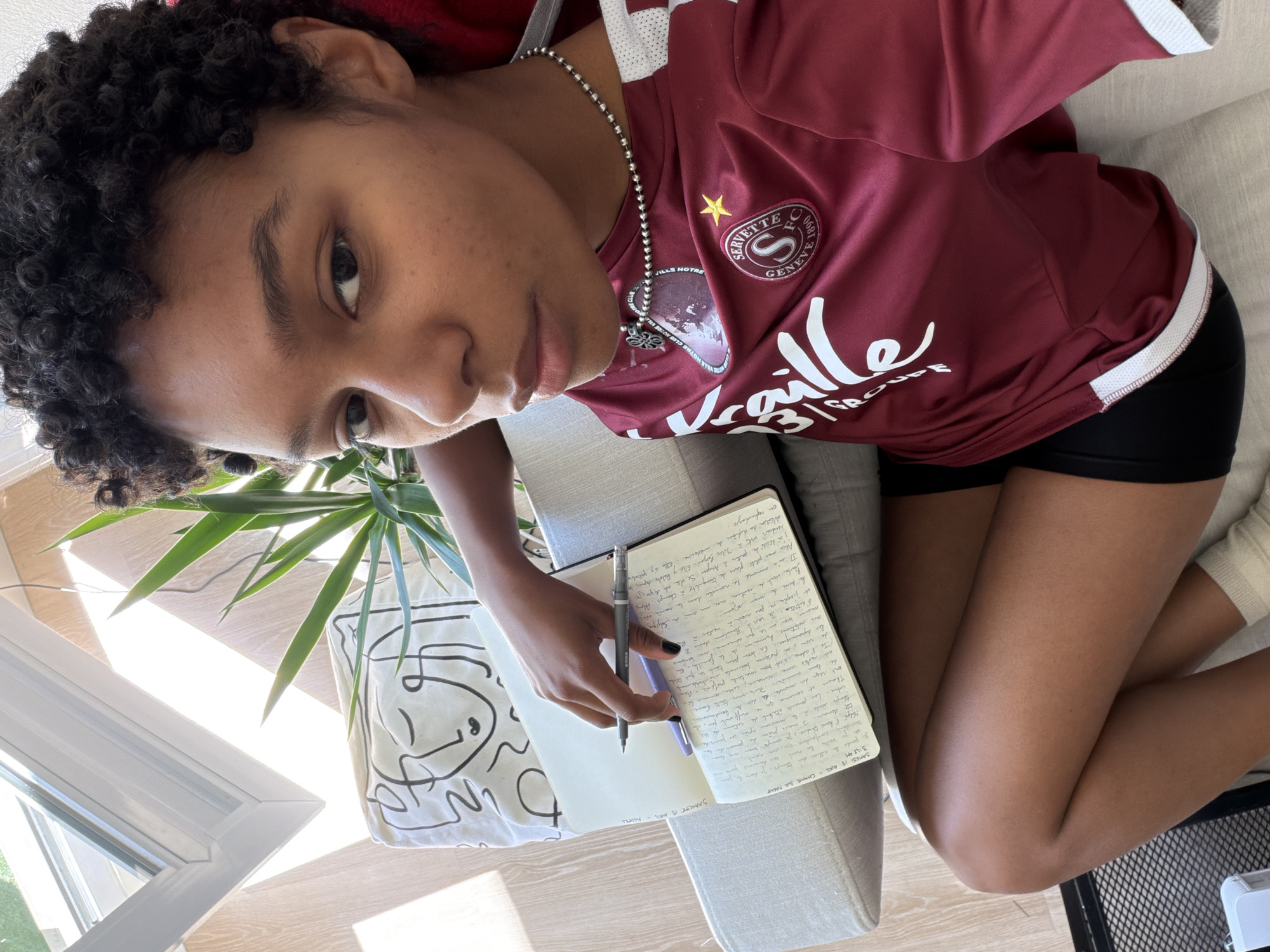 A person with curly dark hair is shown from a high-angle, top-down perspective, apparently writing or about to write in a notebook. The person is wearing a burgundy soccer jersey with a white Servette FC Genève logo. They are also wearing a silver ball chain necklace with a pendant and black shorts. The person's left hand is holding a pen over the open notebook. The notebook contains handwriting in French.
To the left of the notebook, there is a white tote bag with a black line drawing on it and a potted plant. In the background, a window and a slatted wooden ceiling are visible. The handwritten text in the notebook mentions "Angers" and "Julie Bayan," but the location of the image is not explicitly visible in the environment. Therefore, I cannot verify it is in the city of Angers, France.