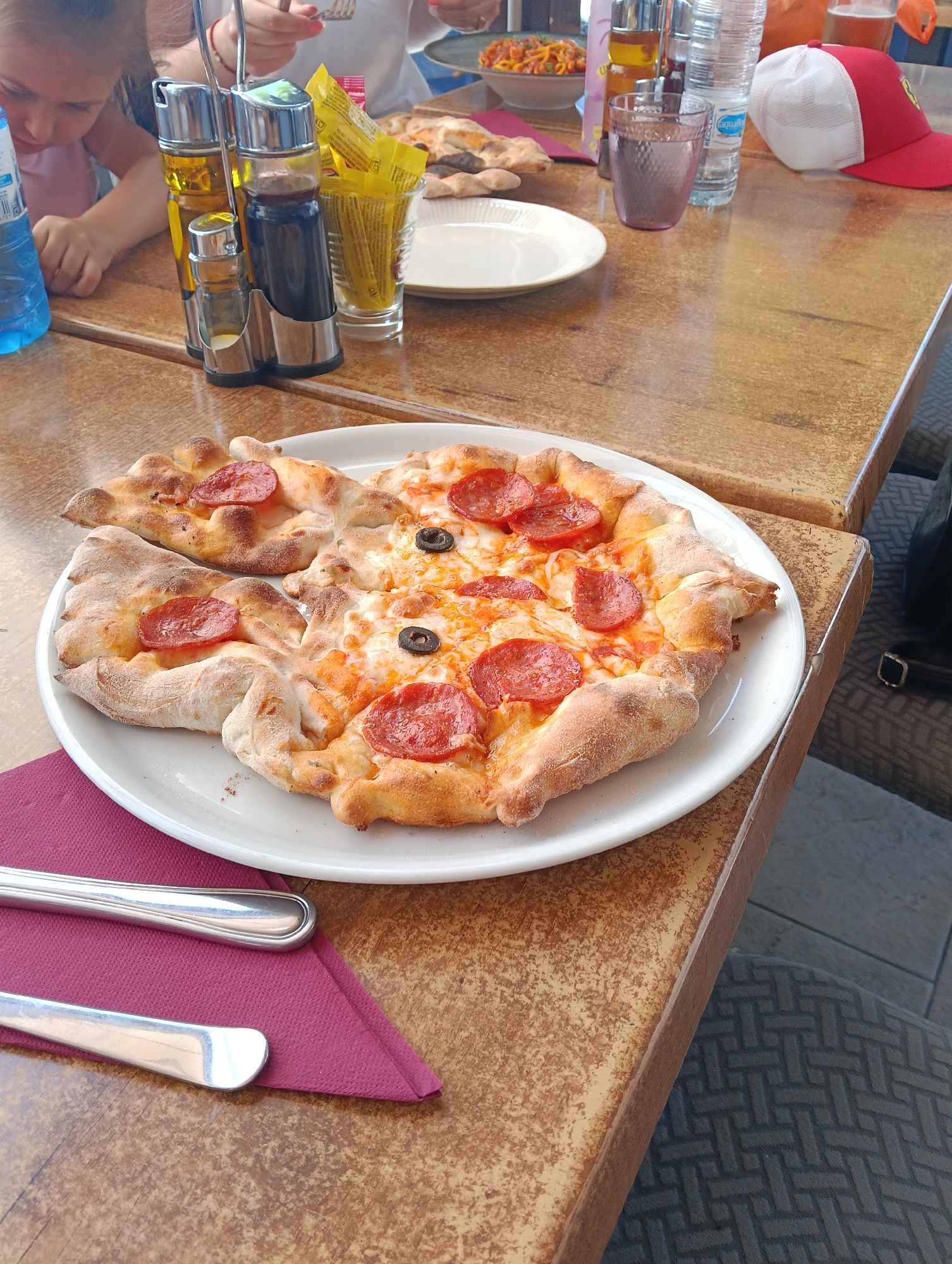 An overhead shot captures an outdoor dining scene at what appears to be a restaurant or cafe in Puerto Del Carmen, Spain. The primary focus is a large, irregularly shaped pizza on a white plate in the foreground. The pizza features a golden-brown crust with some raised, doughy sections, topped with melted cheese, round slices of pepperoni, and a few black olive slices.
To the left of the pizza are a purple napkin and silver cutlery. The table surface is a speckled, light brown material, suggesting a worn or textured finish. In the midground, a chrome caddy holds various condiments: two glass dispensers filled with olive oil and dark liquid (likely balsamic vinegar), and two smaller shakers for salt and pepper. Next to this, a clear glass contains several yellow packets, possibly condiments like mustard or ketchup.
Further back on the table, a blue plastic water bottle with "AQUABONA" visible on its label stands next to a purple-tinted glass filled with water. Another glass, partially obscured, holds a light amber liquid. A red and white baseball cap, possibly bearing a Ferrari logo, rests on the table's edge. In the background, part of a pasta dish in a white bowl with red sauce is visible, along with glimpses of other diners. A child wearing a pink top is seen on the far left, looking down. An adult's hand with pink nail polish is visible further back, holding cutlery.
The scene depicts people enjoying a meal outdoors during daytime, indicated by the bright, natural light. The overall atmosphere suggests a casual and relaxed dining experience. The flooring visible beneath the table features a dark grey, patterned tile or mat.