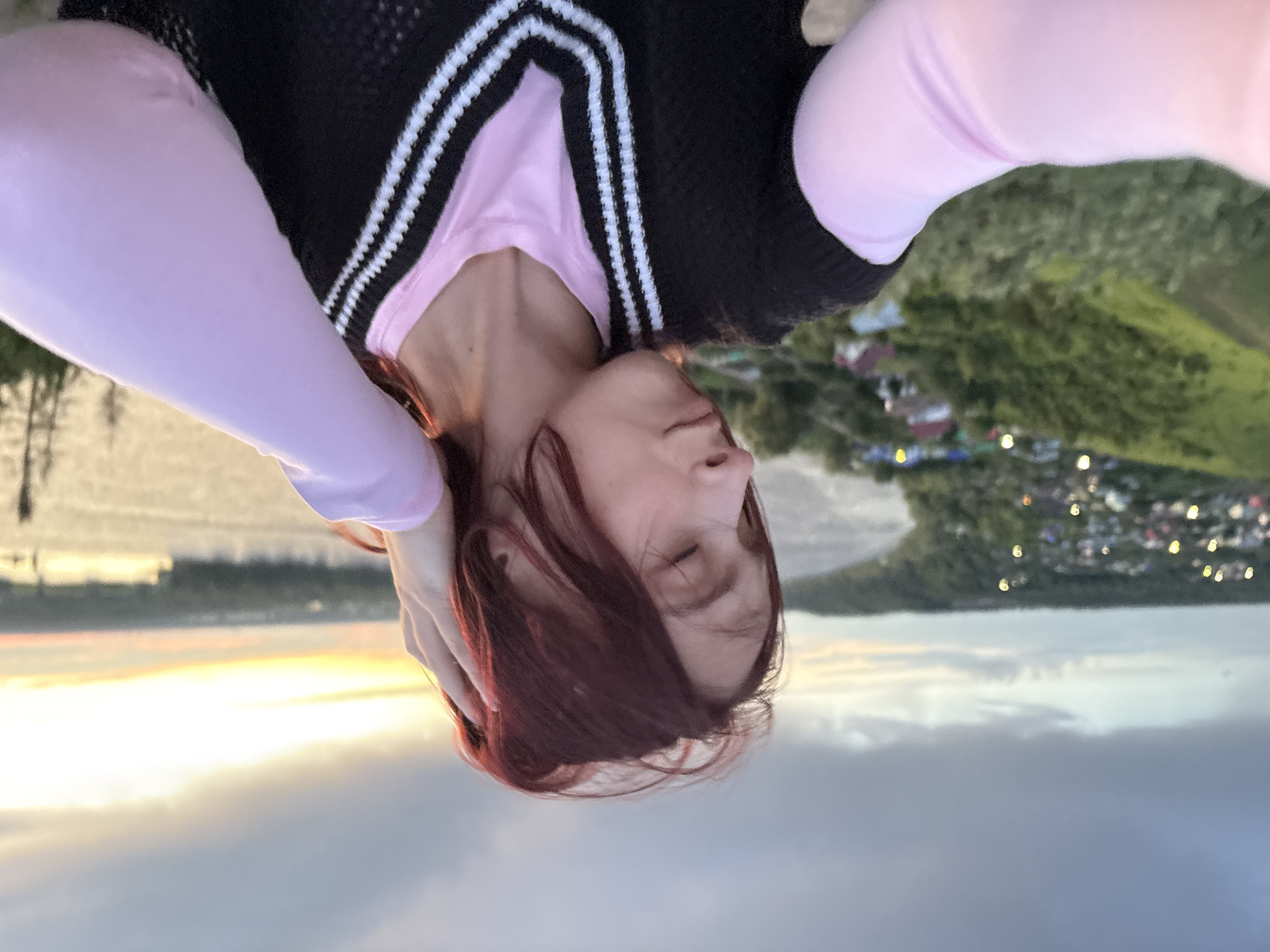 A selfie photograph oriented upside down, taken outdoors with a view of Ольховка, Russia, and what appears to be a body of water reflecting the sky at sunset or sunrise. The subject is a person with reddish-brown hair, their eyes closed, and their hand near their head. They are wearing a pink long-sleeved shirt under a dark vest with white stripes around the V-neck opening. The background includes a village or town along a shoreline and a sky with diffused light.