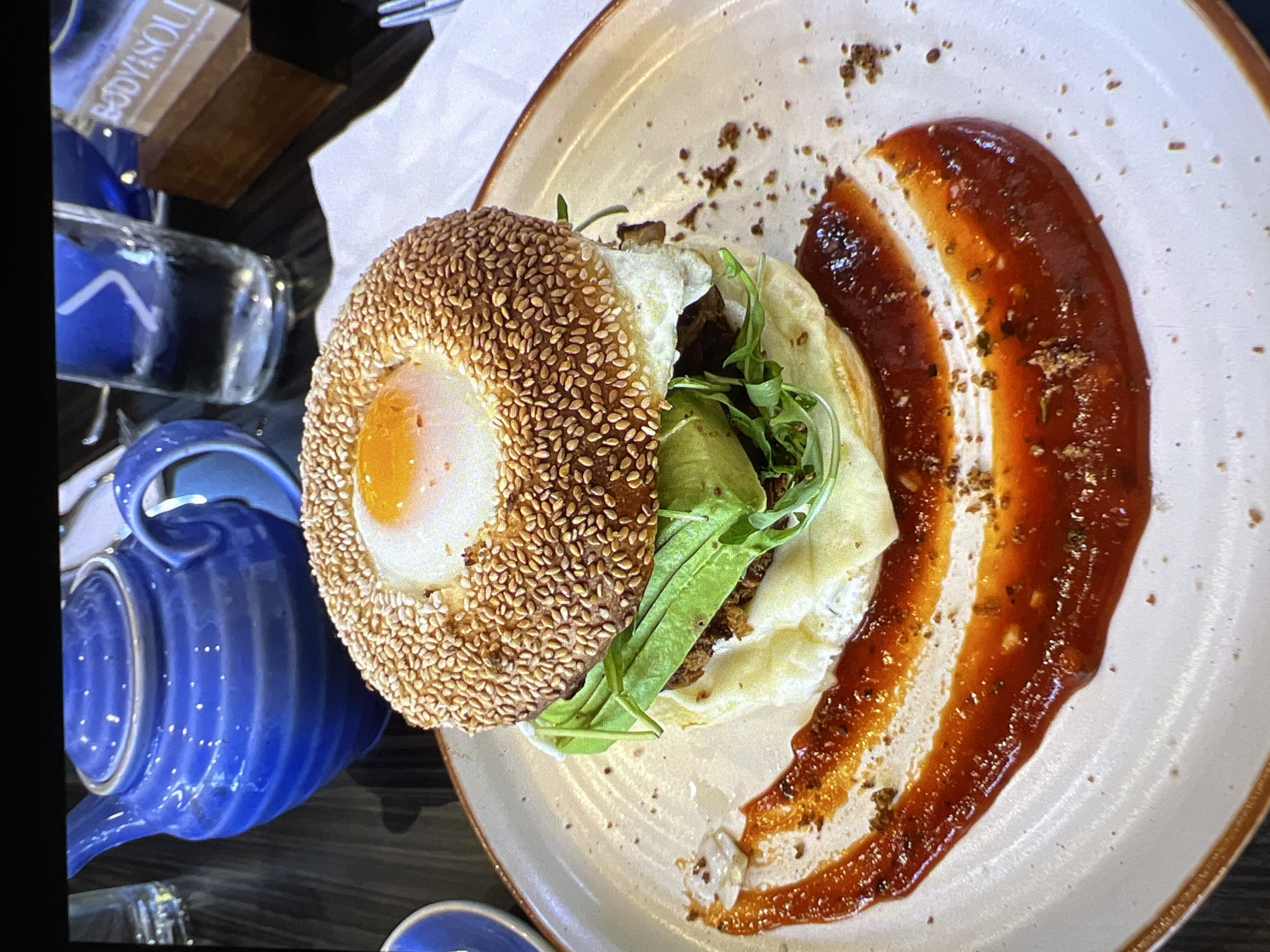 A close-up, eye-level shot captures a breakfast bagel with a fried egg in the center, served on a beige plate with a decorative swipe of red sauce and a sprinkling of herbs. The bagel is generously coated in sesame seeds. Visible through the layers of the bagel are avocado slices and greens. In the blurred background, a vibrant blue teapot sits to the left, and a glass of water is to the right. A wooden sign with the text "BODY+SOUL" is also partially visible. The image does not provide any information about the city of Verona, Italy.