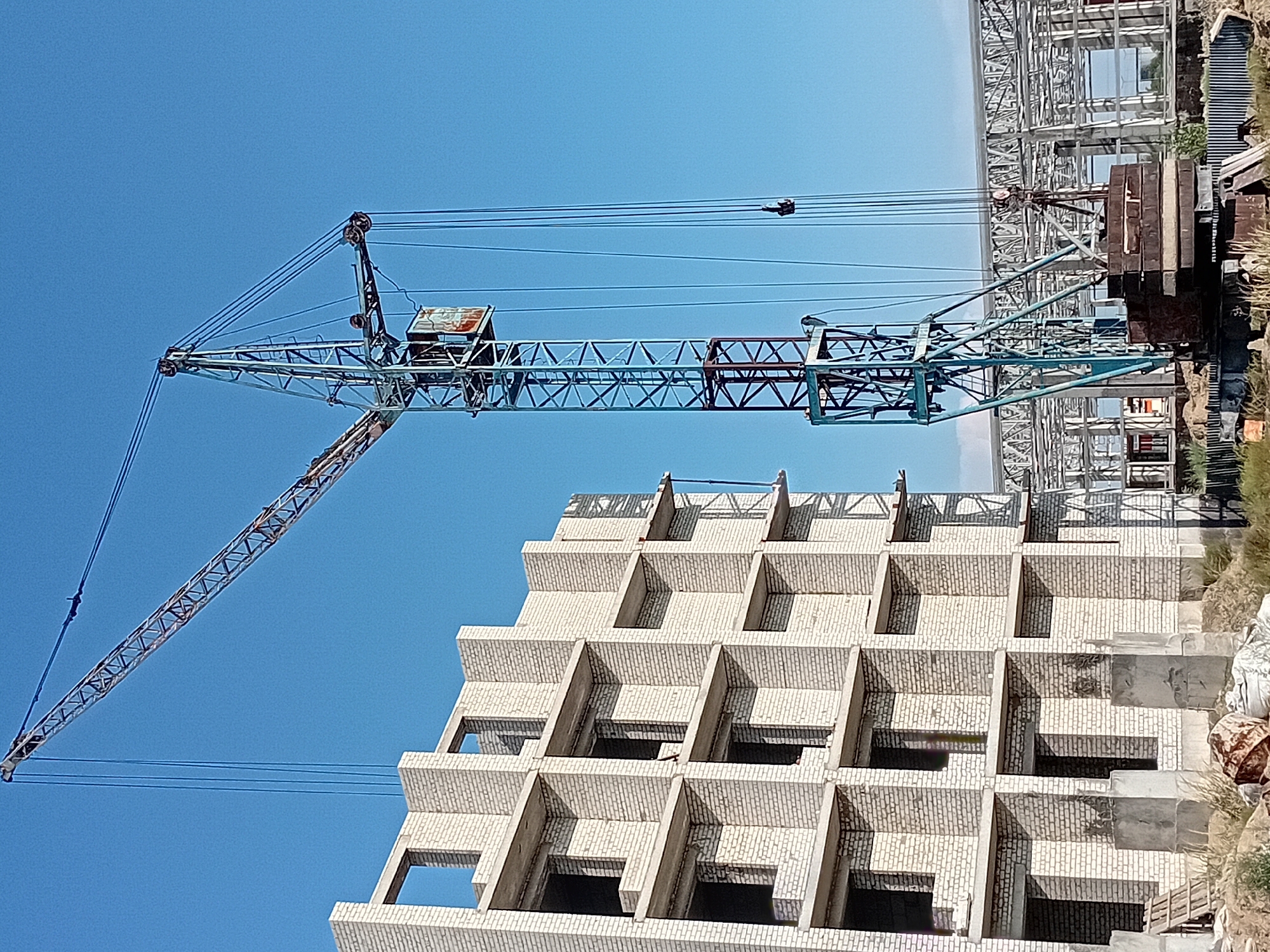 A construction scene is depicted under a clear blue sky in Semey, Kazakhstan. The foreground is occupied by a large, blue construction crane with a red base and a metal cab. The crane is equipped with numerous cables and a hook. To the left of the crane, a multi-story building made of white brick is under construction. The building has a modular design with numerous open window frames and concrete balconies or floor slabs. In the background, the skeletal structure of another large building is visible, suggesting an ongoing construction project. The ground around the construction site is rough and uneven, with some debris and vegetation. The overall impression is one of industrial activity and urban development.