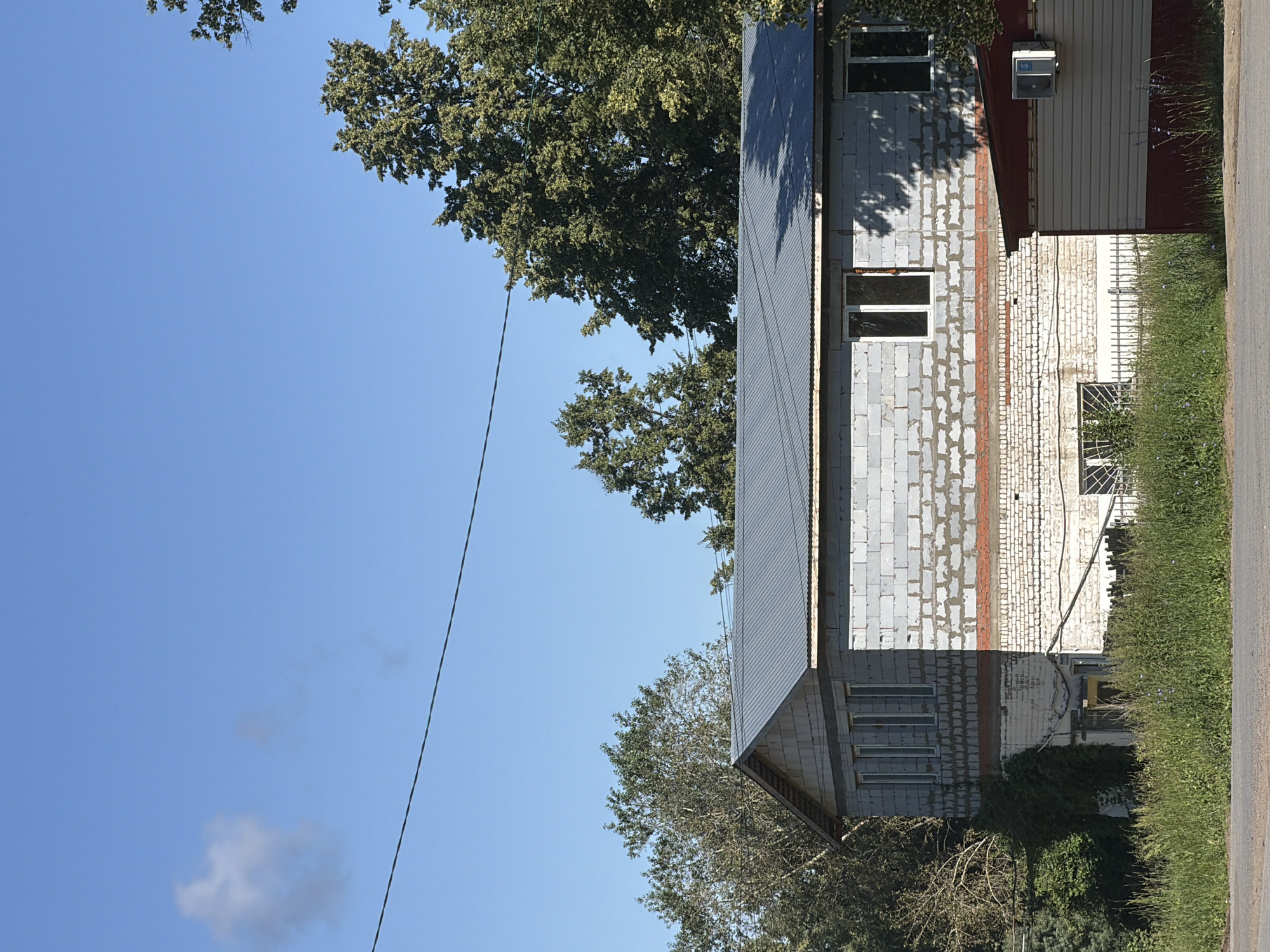 The image captures a two-story building under construction or renovation, constructed from concrete blocks. The building has a gray corrugated metal roof and a lower section faced with white bricks. There are several windows visible, some with white frames and others with dark frames. Adjacent to the main building is a smaller, single-story structure with a red and beige facade, featuring an air conditioning unit mounted on its exterior. The foreground shows a paved road and a stretch of overgrown grass with scattered wildflowers. In the background, lush green trees tower over the buildings, and a clear blue sky with a few wispy clouds completes the scene. The image appears to be taken in Chaikovsky, Russia, a city known for its industrial and residential areas surrounded by nature.