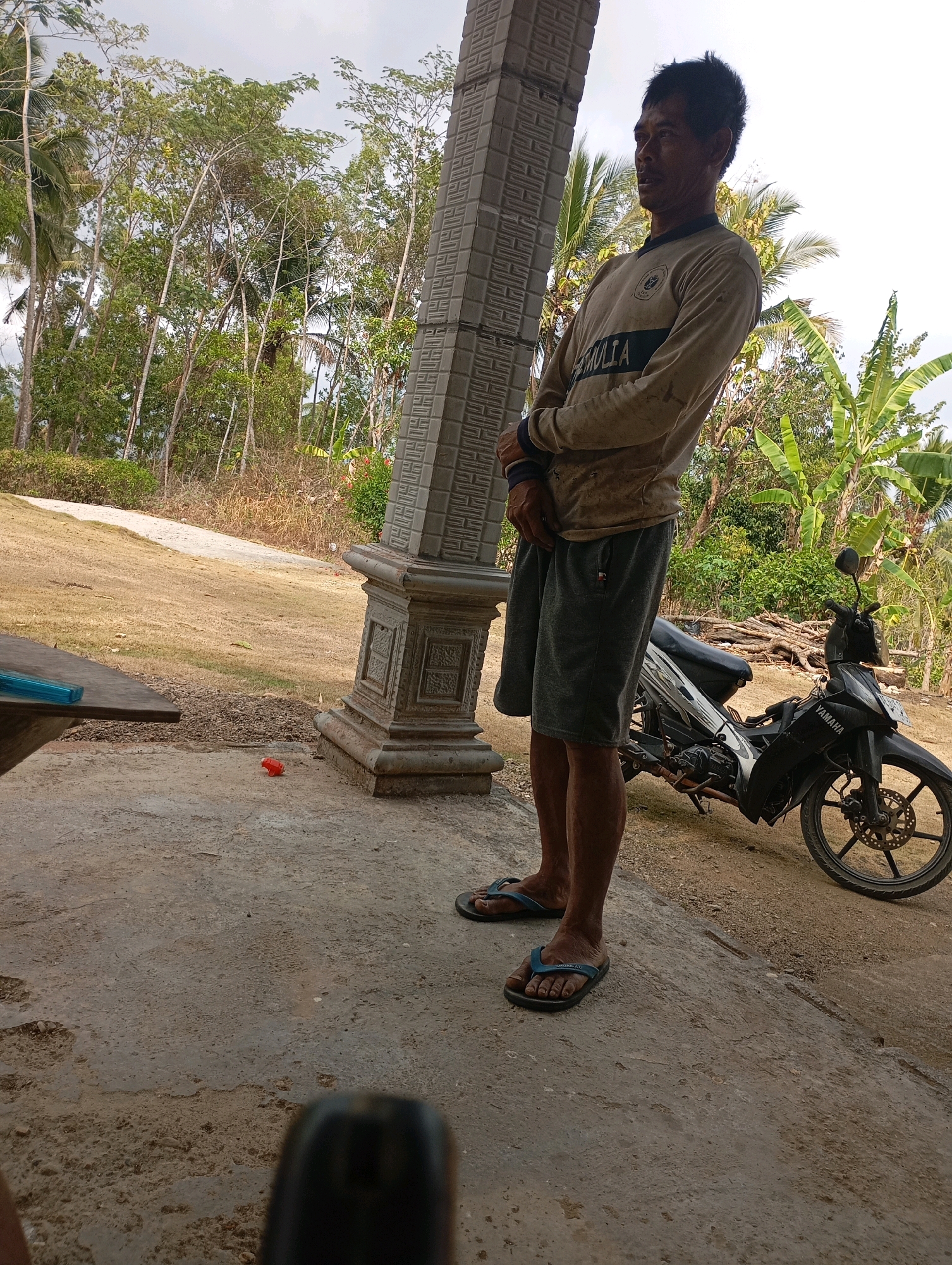 The image depicts a man standing outdoors, in what appears to be a rural or semi-rural setting in Ketepung, Indonesia. He is positioned to the right of a decorative concrete pillar and facing slightly to his right. The man is wearing a long-sleeved, light-colored shirt with some dark markings and grey shorts. He has dark hair and a mustache. On the ground near him, to the left, is a black motorcycle with the "YAMAHA" logo visible. The background consists of trees, some of which are palm trees, and lush vegetation, suggesting a tropical environment. The ground is a mix of concrete and dirt. A portion of a wooden table with a blue object on it is visible in the lower left corner. The sky is overcast.