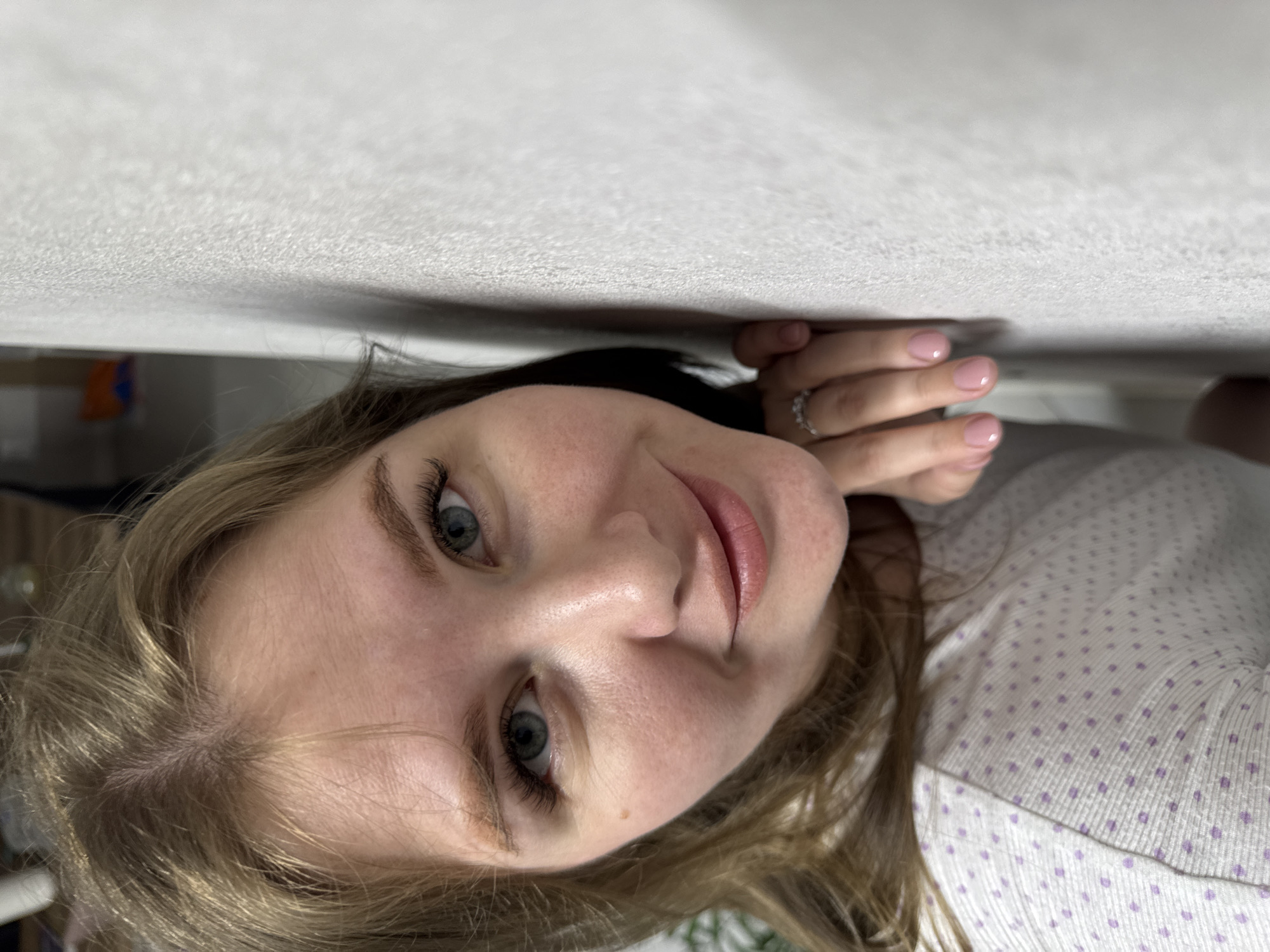 The scene depicts a close-up, upward-angle portrait of a fair-skinned female located in Gornyi (Dvurechenskoe S/P), Russia. The subject has blonde or light brown hair with a central part, blue eyes with dark eyelashes, light-colored eyebrows, and full lips, displaying a subtle smile directed at the camera. A singular mole is present on her left cheek, below the eye. She is wearing a light-colored, possibly white, top with small, repeating purple polka dots. Her right hand is visible, resting against a light grey, textured wall that dominates the right portion of the frame. The hand features manicured fingernails with light pink polish, and a ring with multiple small, clear stones is worn on her right ring finger. The background on the left is out of focus, revealing indistinct interior elements and a blurred hint of green foliage in the far left.