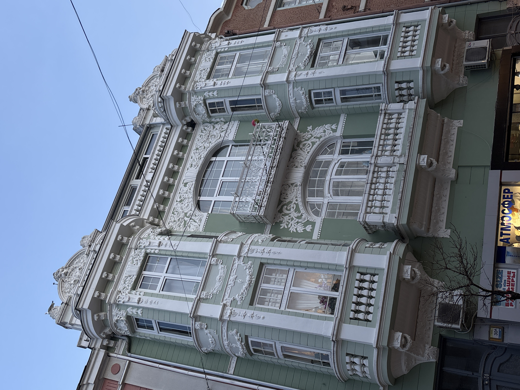 The image displays the ornate facade of a multi-story building located in Varna, Bulgaria. The structure is painted a pale mint green, extensively adorned with white decorative stucco featuring intricate floral and scroll patterns, along with classical elements. The building incorporates two prominent bay windows, each with white balustrade railings at its base, projecting from the upper floors. A central balcony with white wrought-iron railings is present on an upper level, where a small colorful object, possibly a flower, is visible. Another small pink object is situated near the right bay window.
The fenestration includes numerous white-framed windows, some rectangular and others with arched tops. The roofline is characterized by elaborate pediments, with a white bird perched atop the leftmost one. Thin black utility wires traverse the clear blue sky visible above the building.
The ground floor features a commercial storefront with visible signage displaying "BIG SIZE" and "АТМОСФЕР" (ATMOSFER) in Cyrillic. Two white external air conditioning units are mounted on the lower facade. A bare-branched tree is positioned in front of the lower left section of the building, near a dark door marked with "30" and "30A". Portions of adjacent buildings, one with a pale pink/grey facade on the left and one with red brick on the right, are partially visible. No individuals are depicted in the scene.