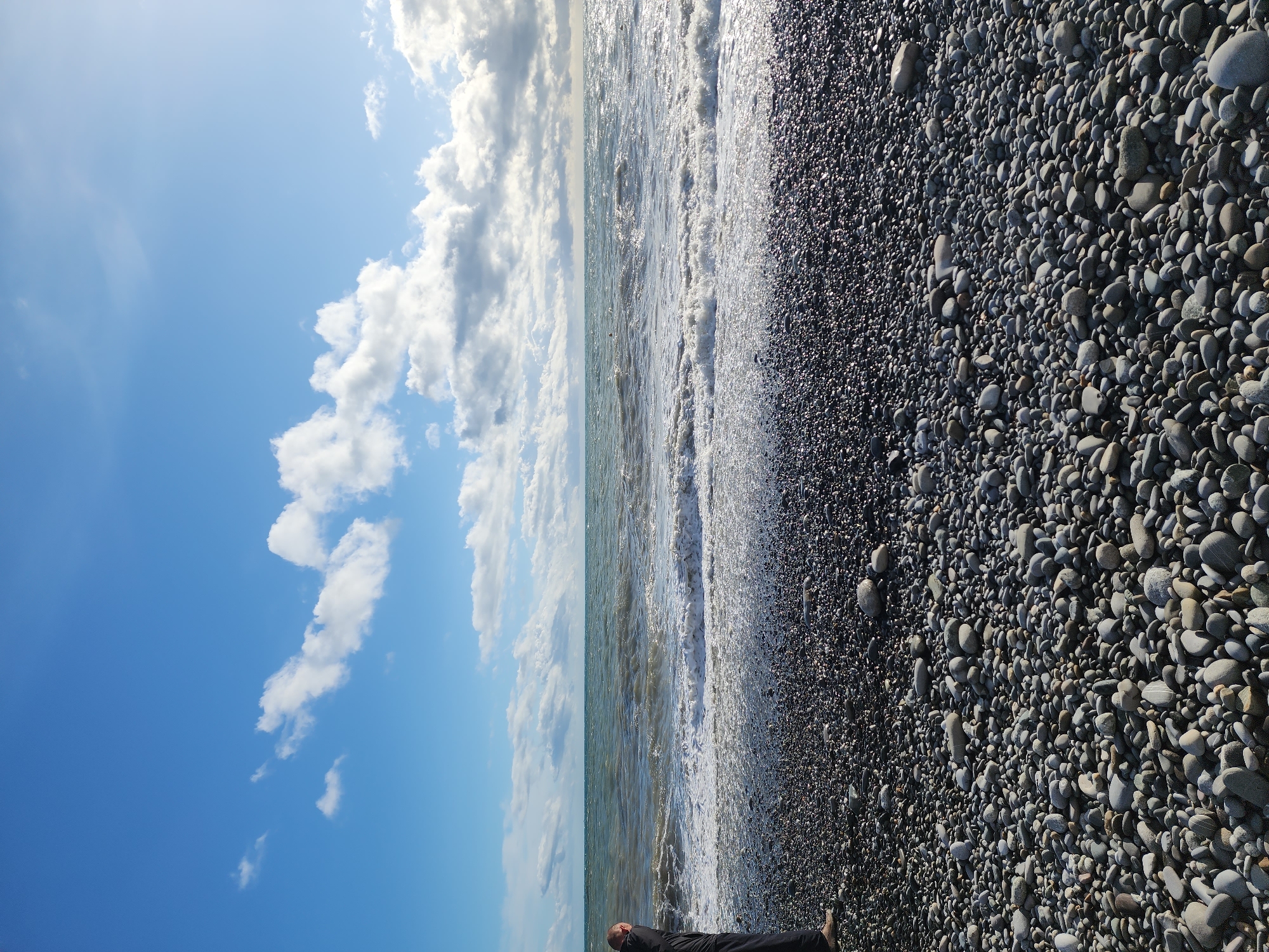 A beach in Sochi, Russia, is captured on a sunny day with scattered clouds in the bright blue sky. The foreground is a pebble beach covered in smooth, rounded stones varying in shades from light gray to almost black. The sea meets the shoreline with small waves, creating a foamy white edge between the water and the pebbles. A person, dressed in dark attire, lies near the bottom of the frame, partially visible.